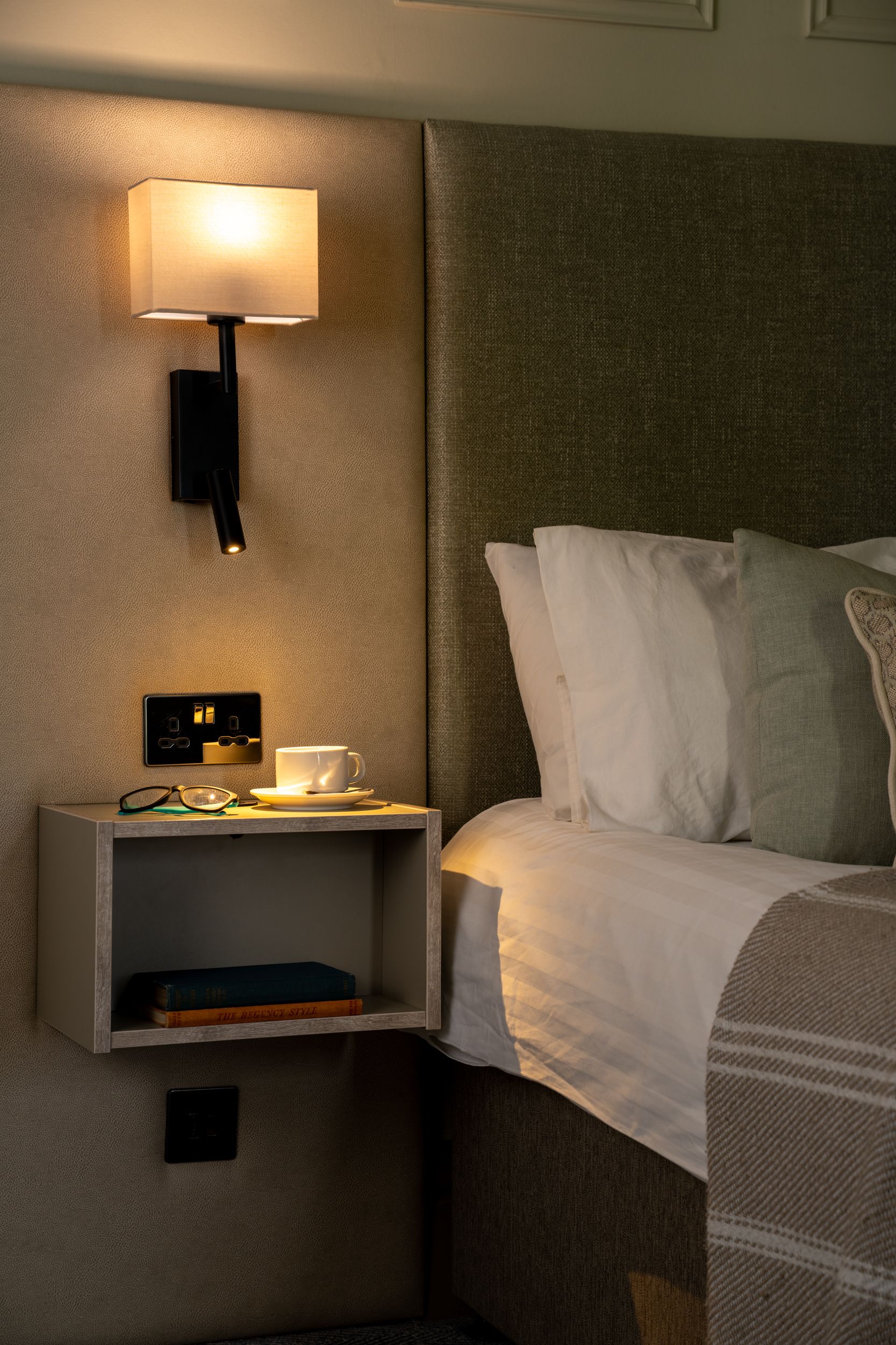 Modern hotel room nightstand with a cup and glasses, wall lamp above, and neatly made bed.