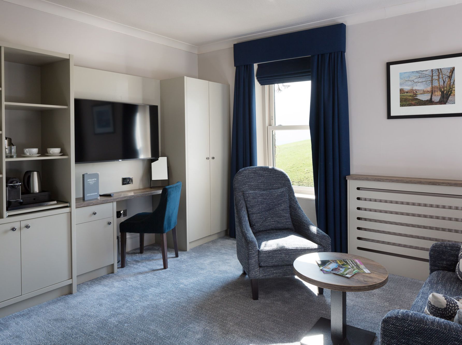 Modern hotel room with lounge chair, TV, desk, and window with blue curtains