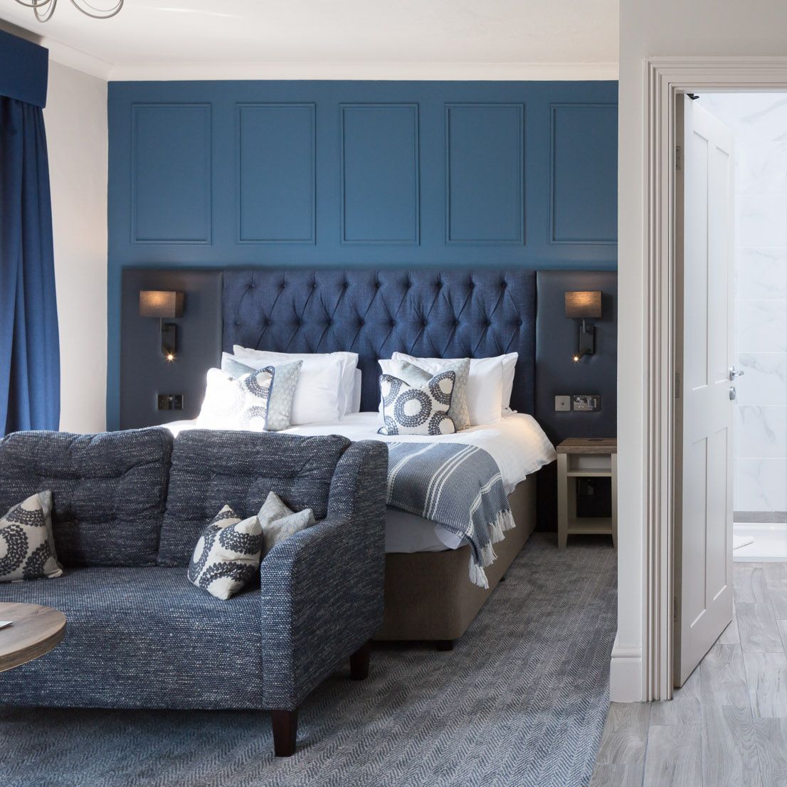 Modern hotel room with blue accents, visible bed, sofa, freestanding bathtub, and a bathroom entrance.