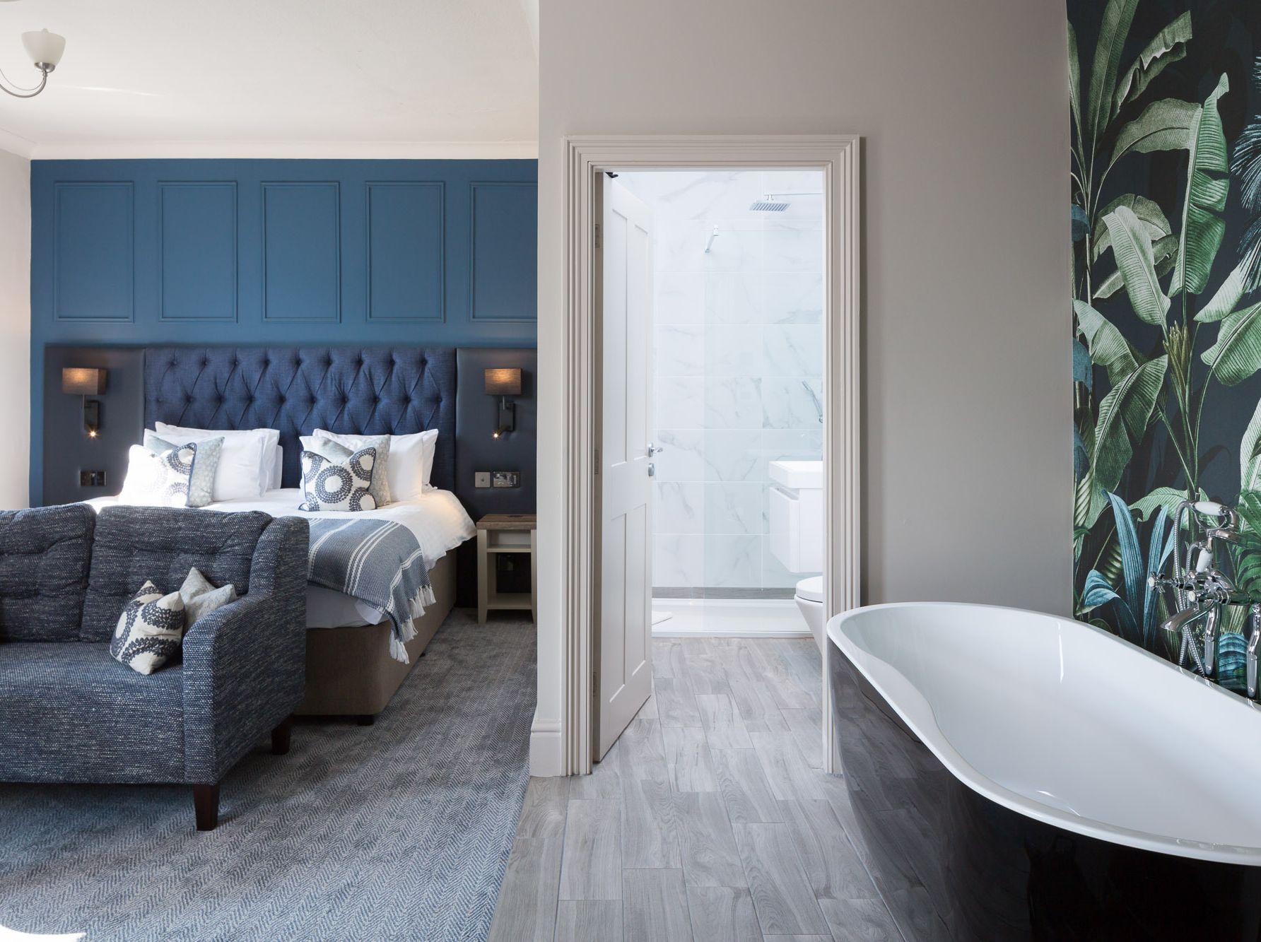 Modern hotel room with blue accents, visible bed, sofa, freestanding bathtub, and a bathroom entrance.