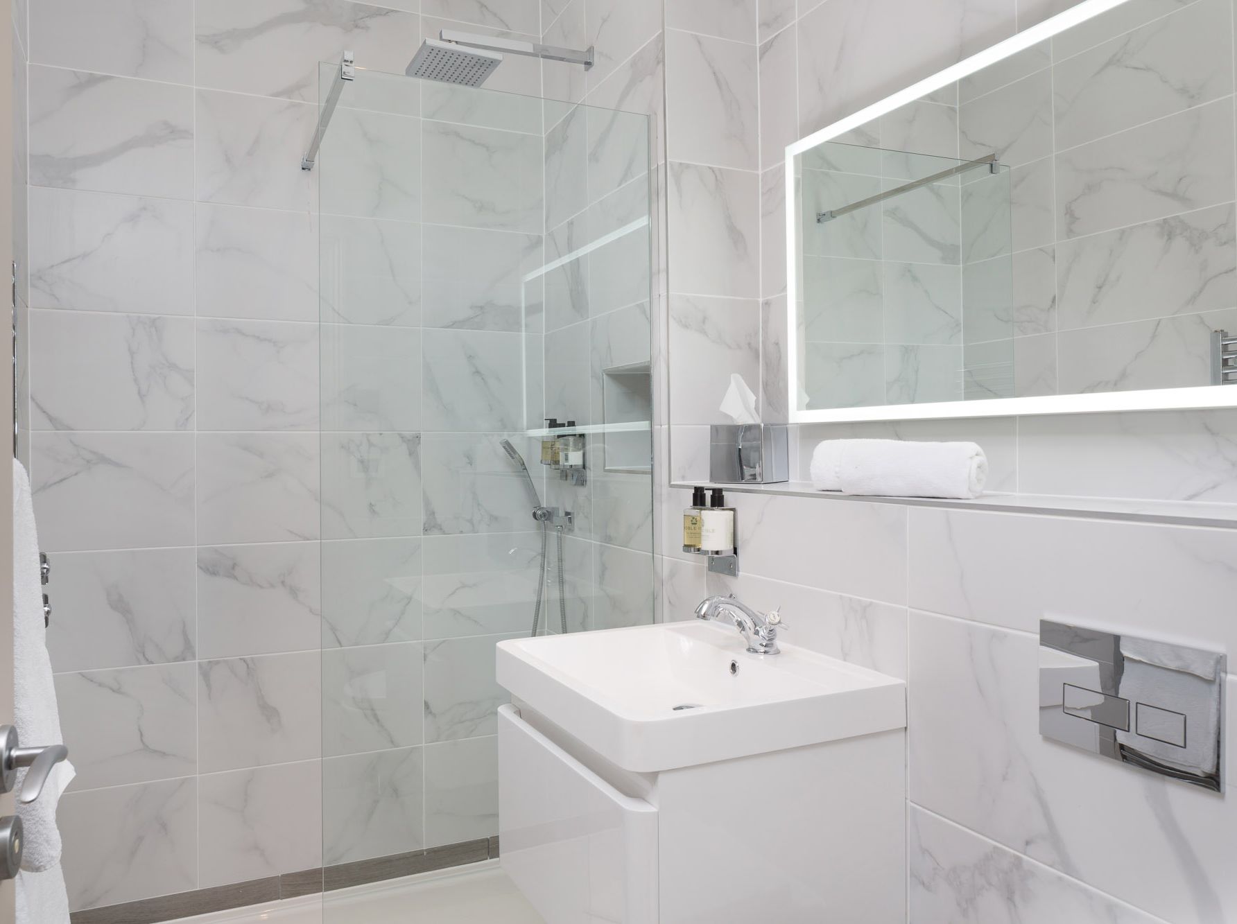Modern bathroom with marble tiles, glass shower, illuminated mirror, and white sink.