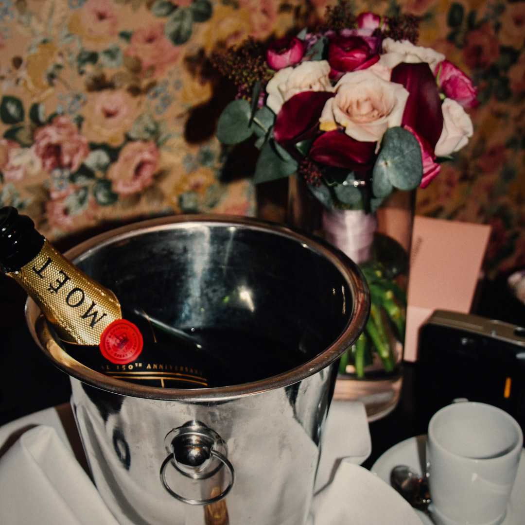 A bottle of Moët champagne chilling in an ice bucket next to a vase of roses and a cup on a saucer.