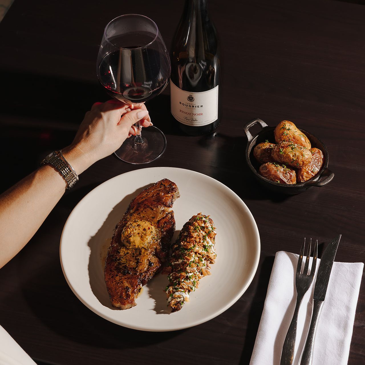 A meal with steak, twice baked potato, roasted potatoes, a glass of red wine, and a bottle of Pinot Noir on a dark table.