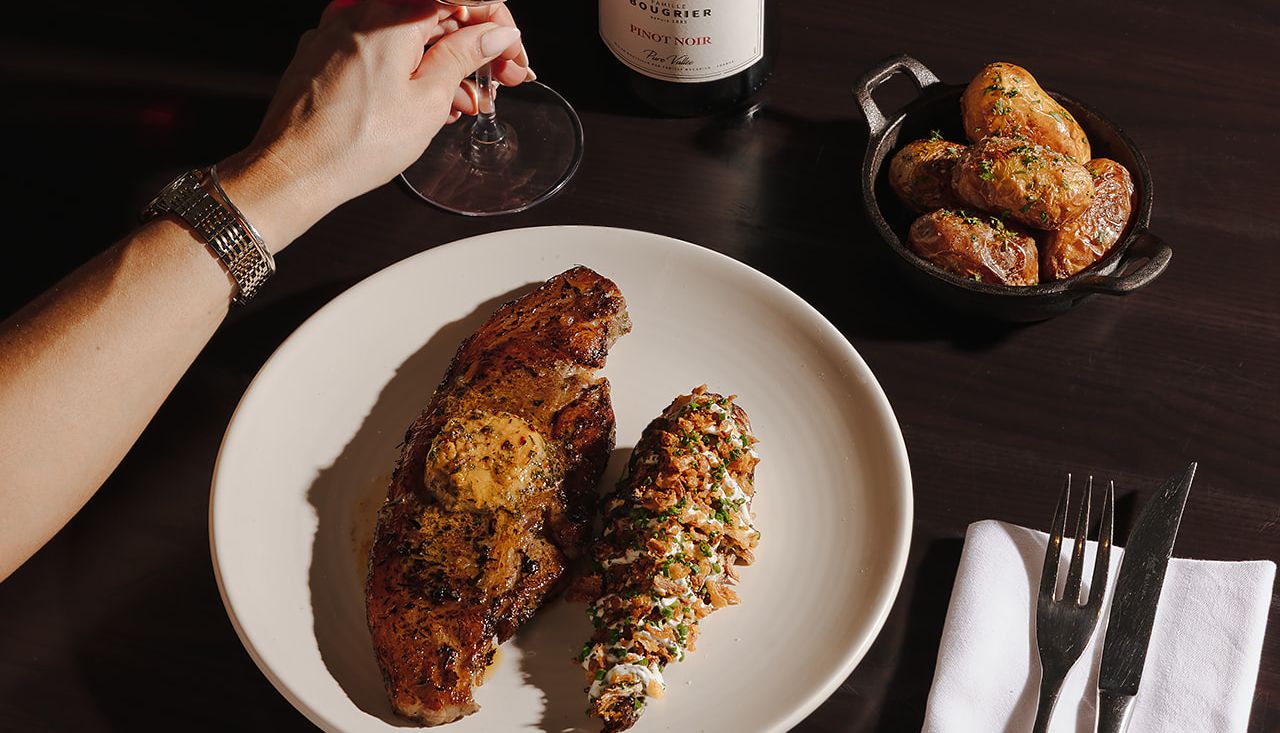 A meal with steak, twice baked potato, roasted potatoes, a glass of red wine, and a bottle of Pinot Noir on a dark table.
