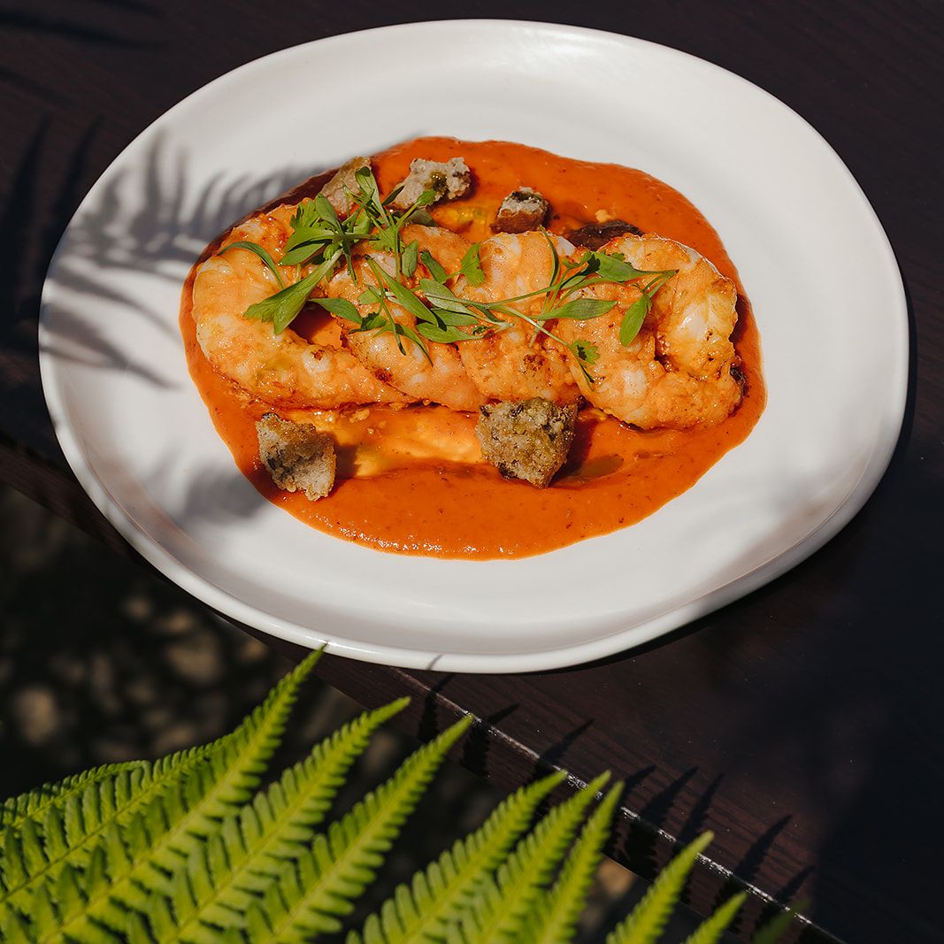 A plate of shrimp in a rich orange sauce, garnished with herbs and croutons, set on a dark table with a bright green fern in the foreground.