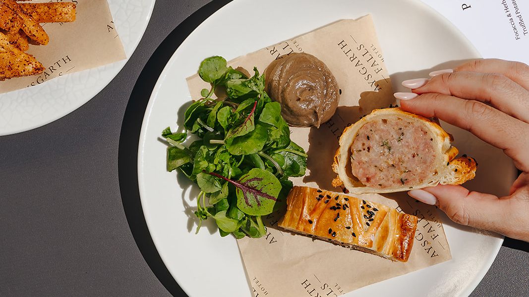 A selection of appetizers including a bowl of mixed olives, a glass of rosé wine, and a plate with a sausage roll, greens, and pâté.