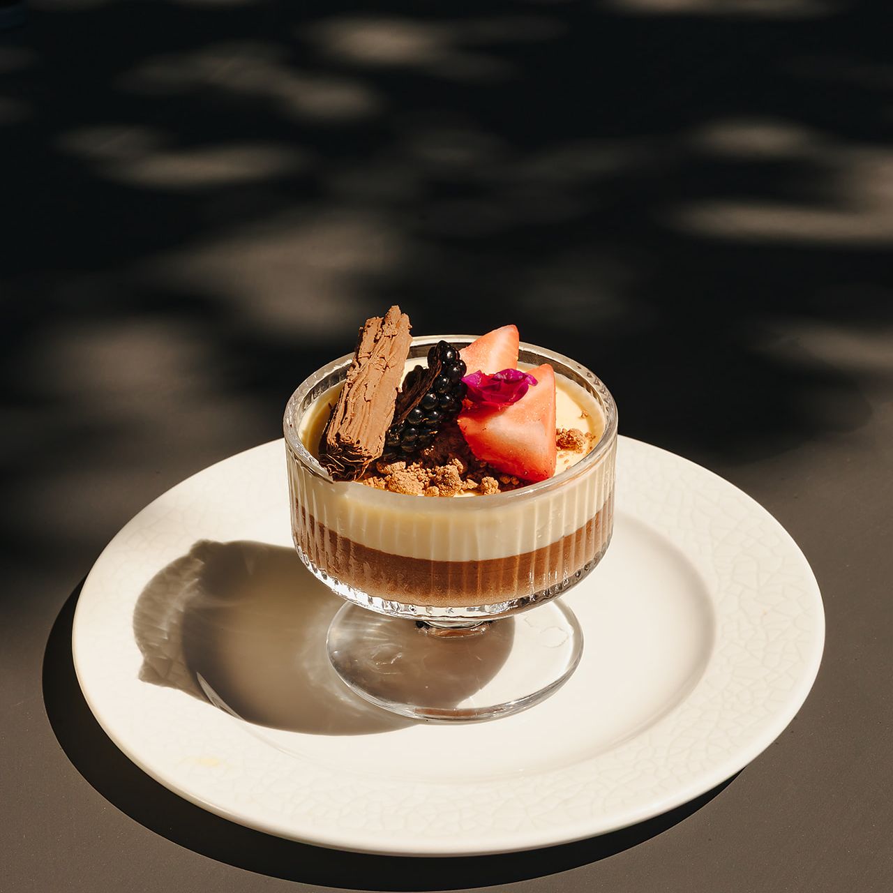 Glass dessert cup filled with layered mousse, topped with fresh fruit, chocolate flake, and crumble, served on a white plate.
