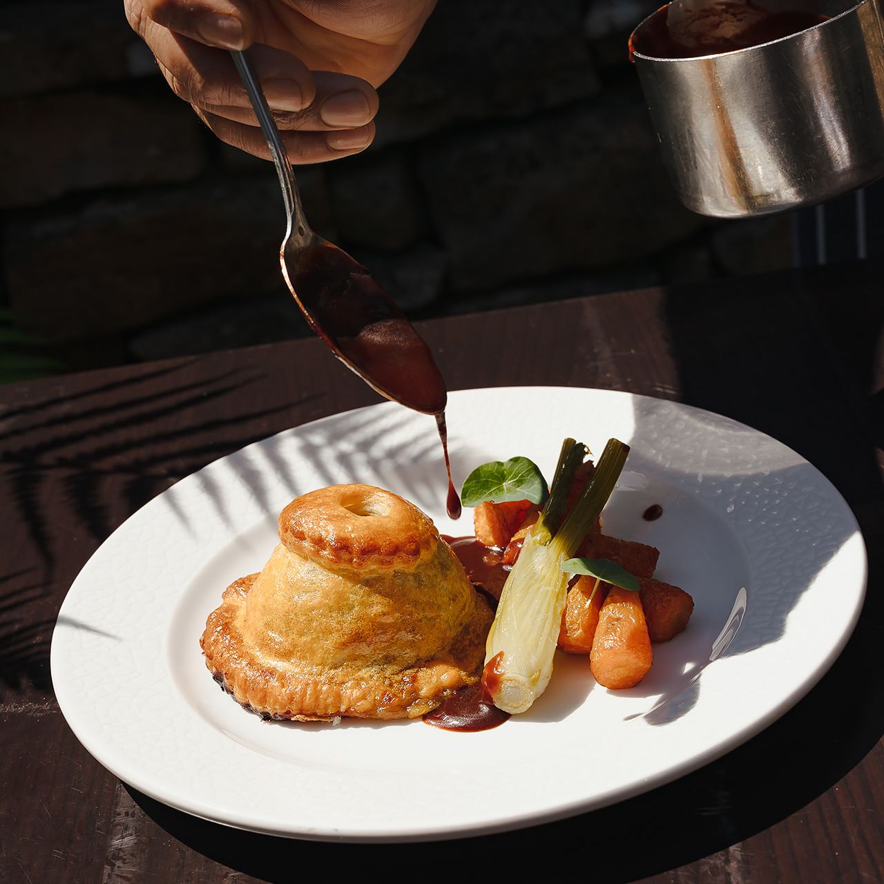 Close-up of a hand pouring sauce over a small savory pie with vegetables on a white plate.