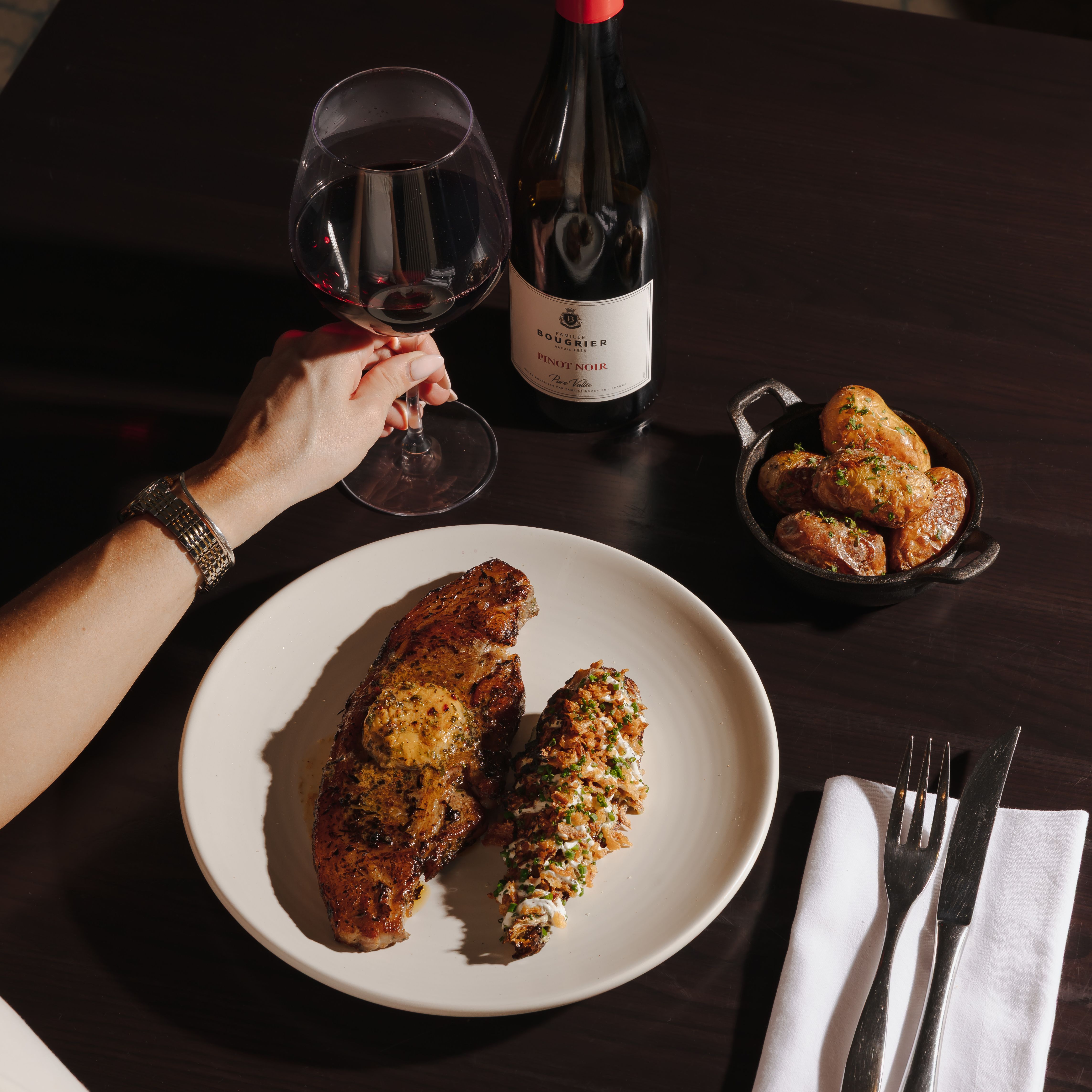 A person holding a glass of red wine beside a plate of steak with a stuffed potato, roasted potatoes, and a bottle of Pinot Noir on a dark table.