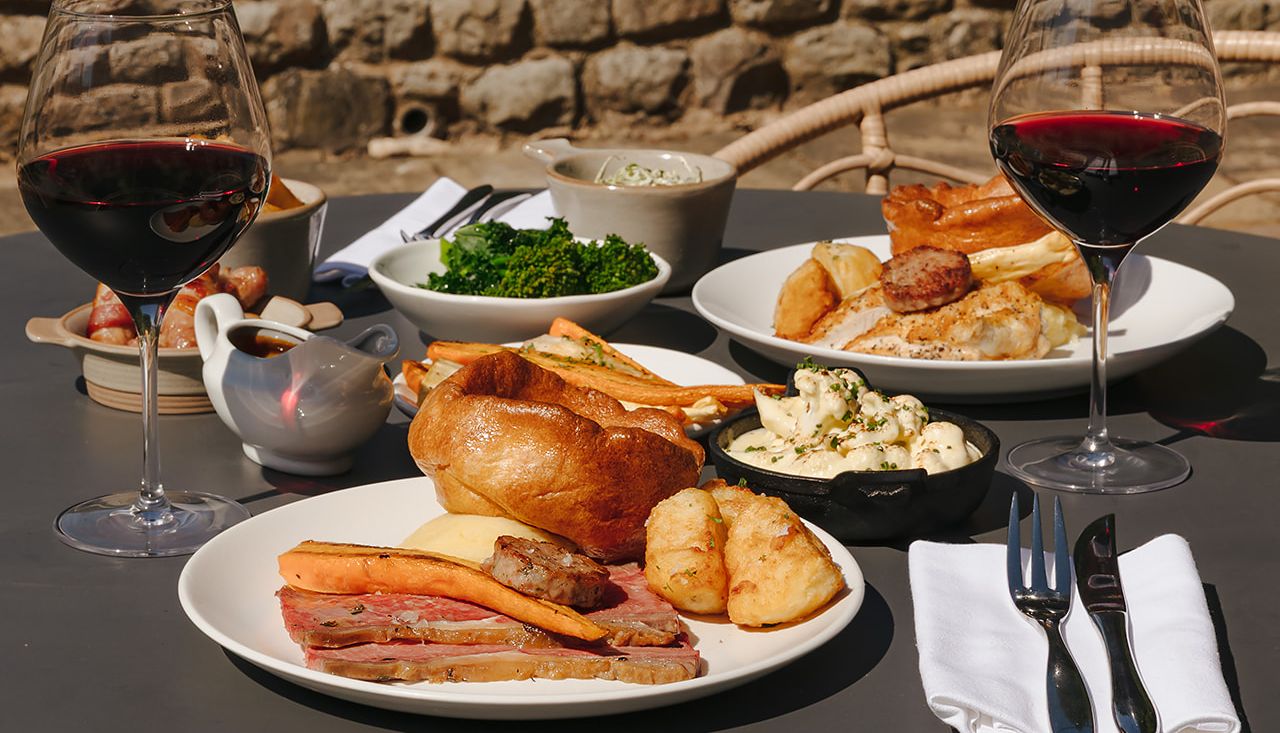 Two plates of traditional British roast dinners with Yorkshire pudding, vegetables, and glasses of red wine on an outdoor table.