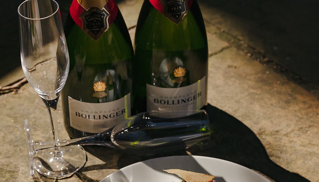 Two bottles of Bollinger champagne, two champagne flutes, and a gourmet appetizer on a white plate.