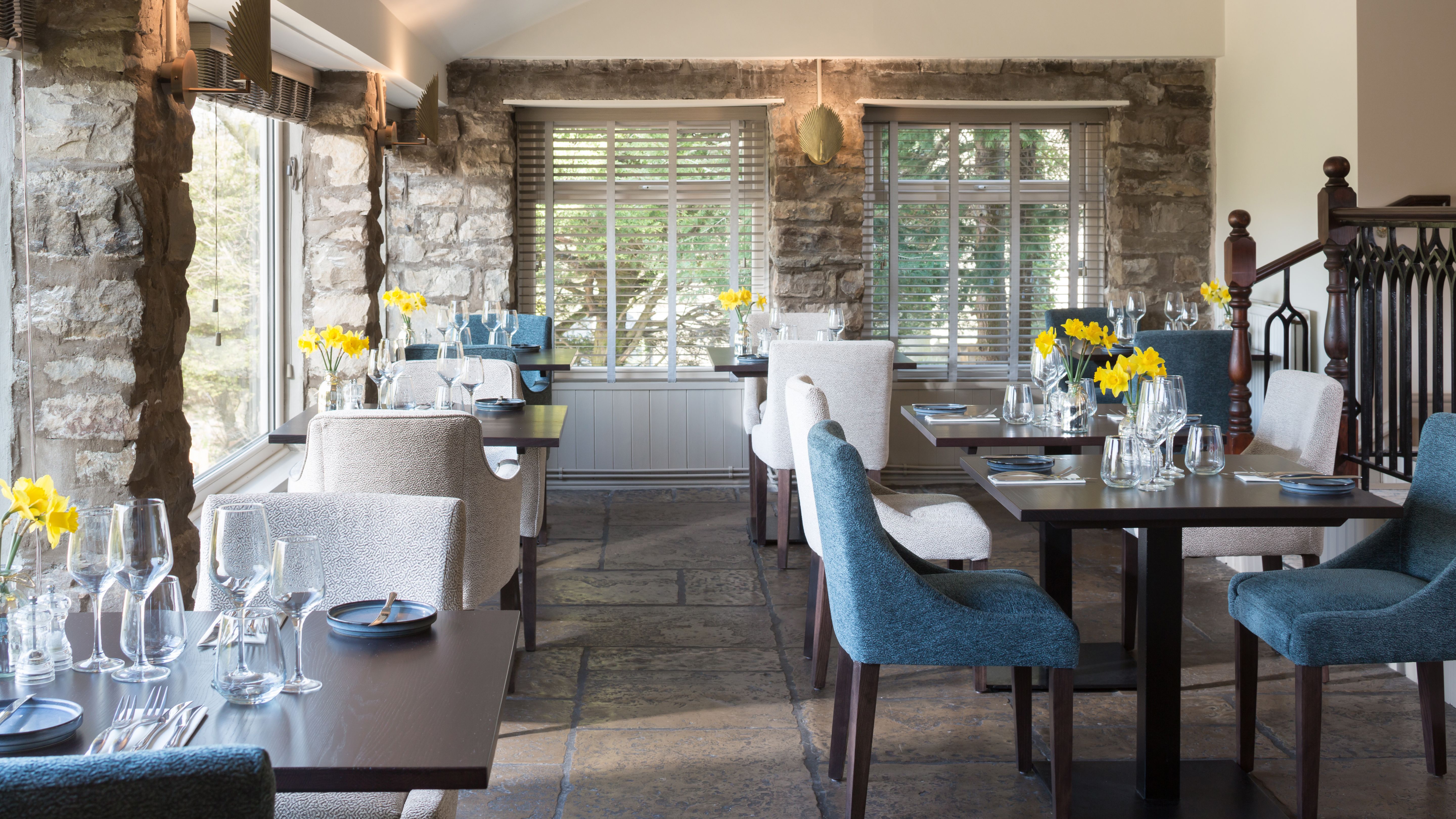 Cozy restaurant interior with stone walls, large windows, and tables set with glassware and yellow flowers.