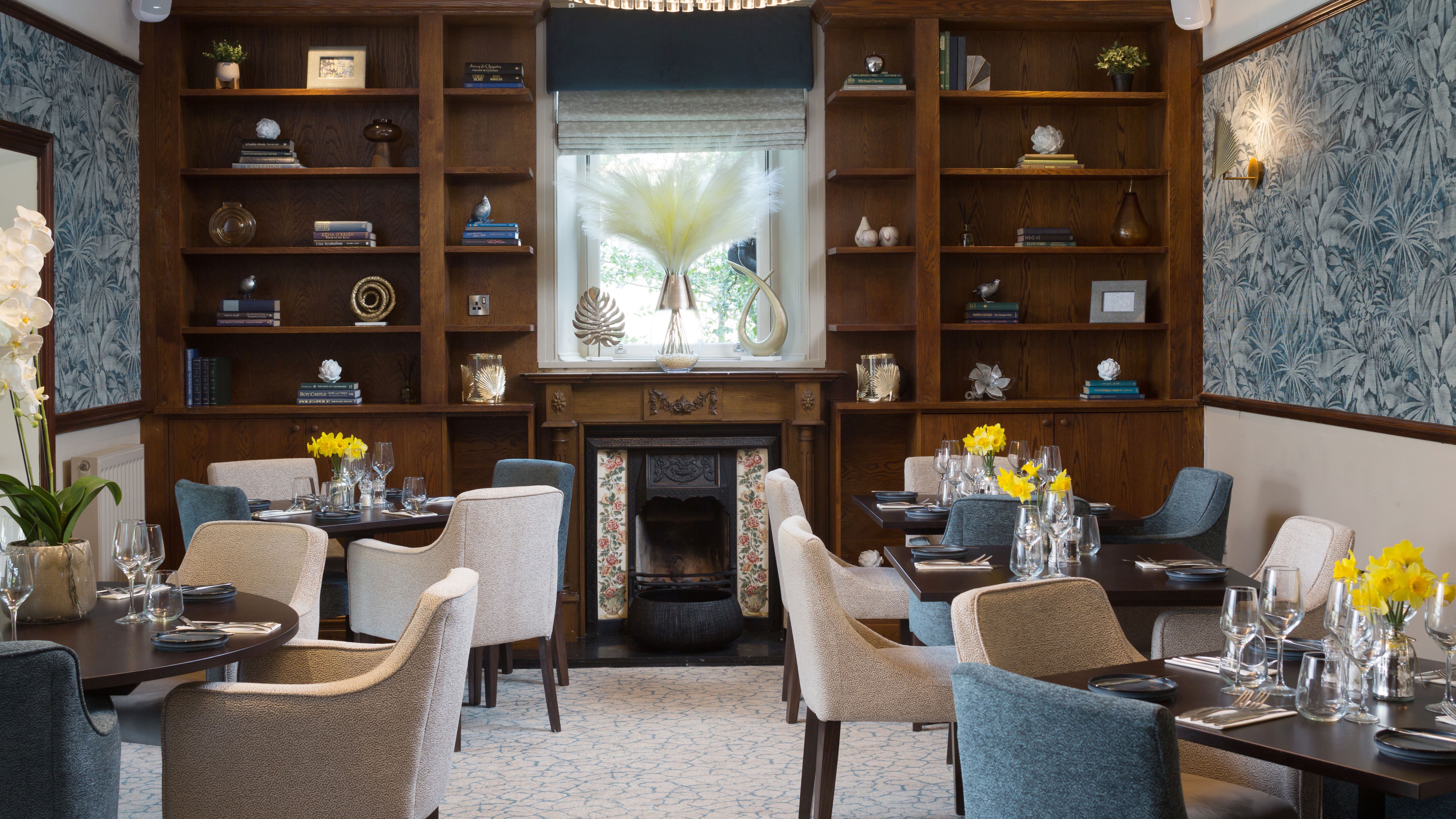 Elegant restaurant dining area with blue and cream chairs, wooden bookshelves, and a decorative fireplace.