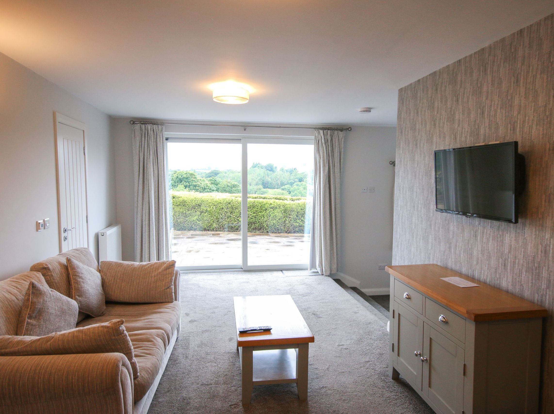 Modern living room with beige sofa, wall-mounted TV, and large sliding glass doors opening to a patio with greenery outside.