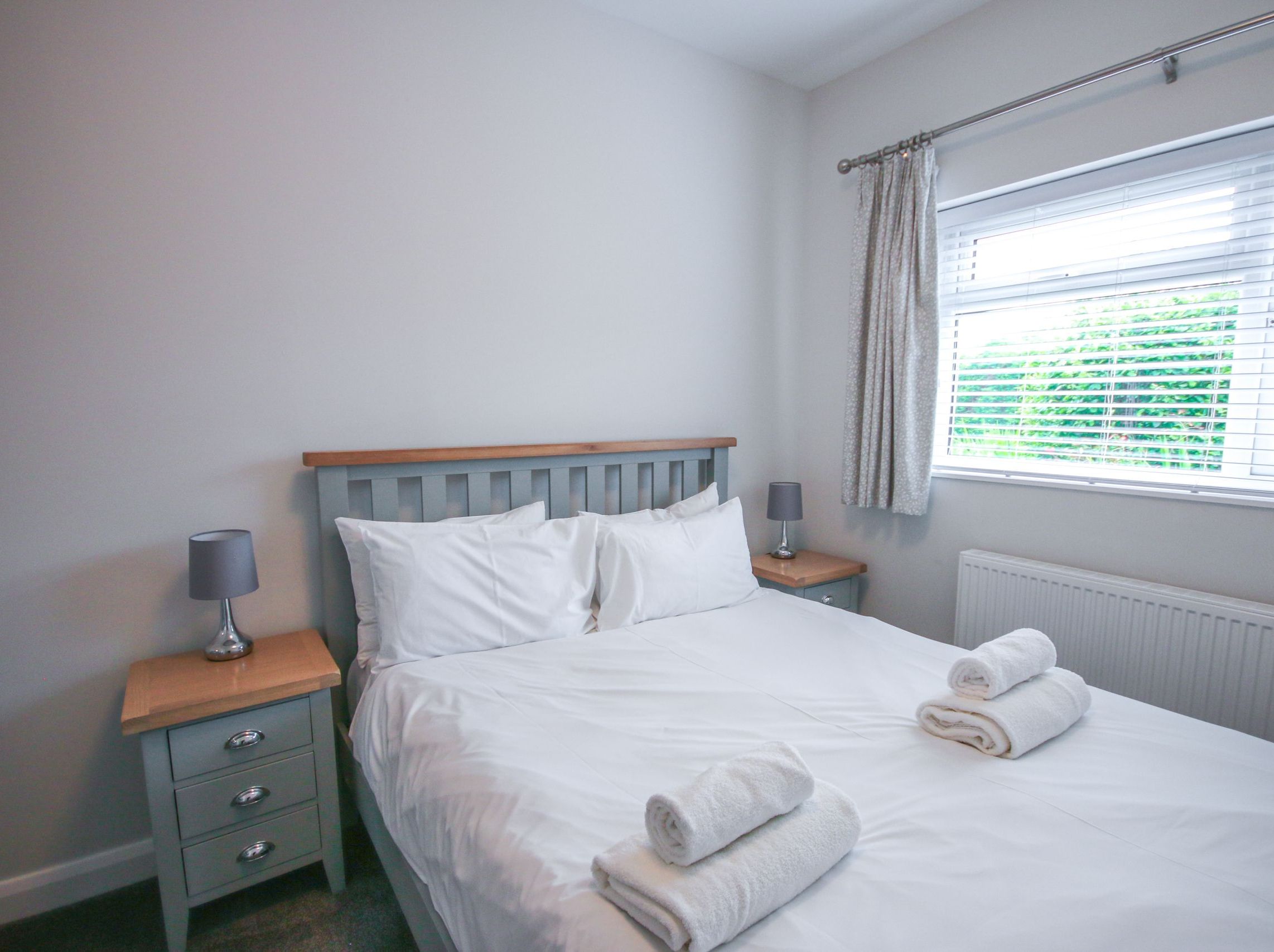 Neatly arranged bedroom with a double bed, white linens, bedside tables, lamps, and a window with blinds.