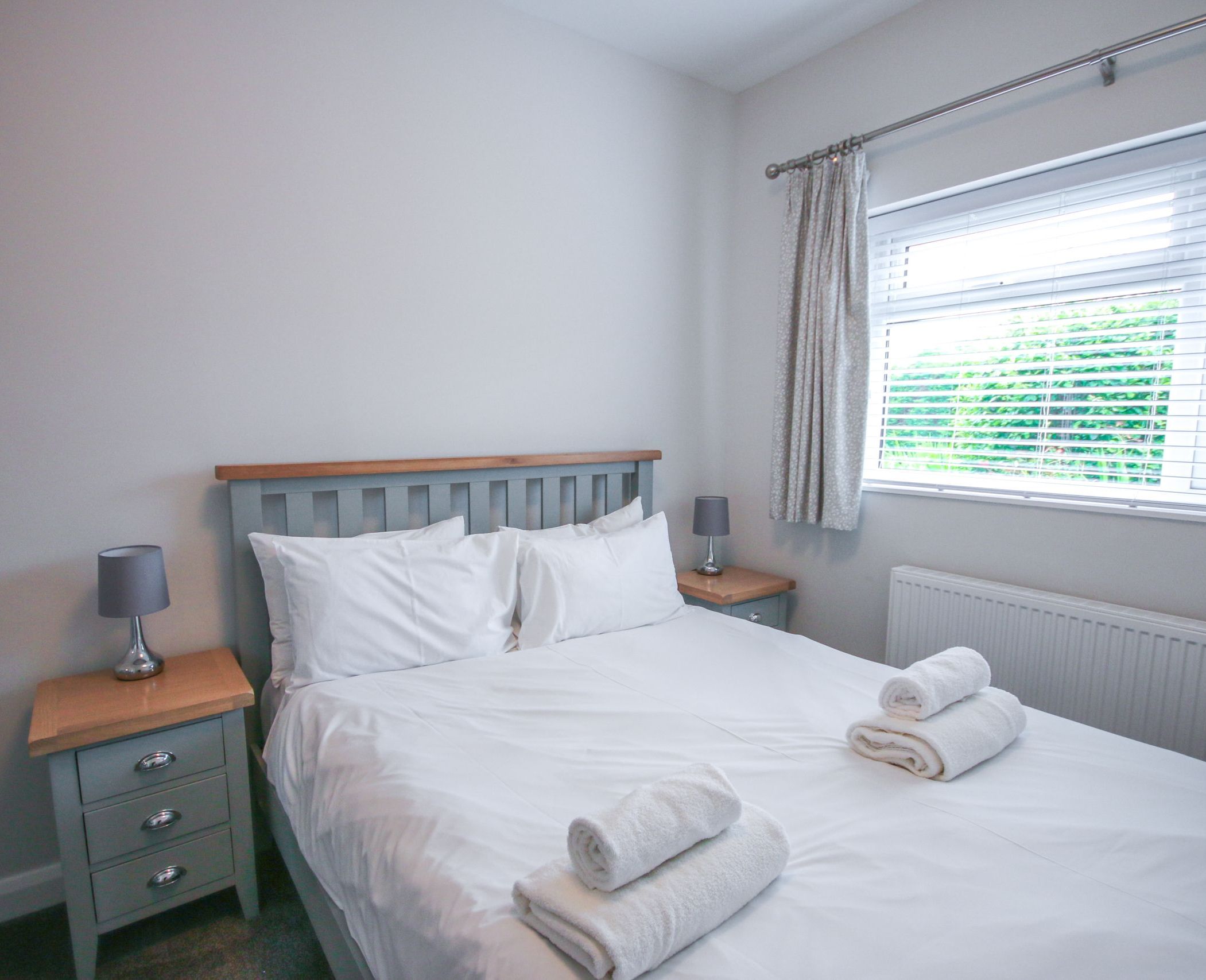Neatly arranged bedroom with a double bed, white linens, bedside tables, lamps, and a window with blinds.
