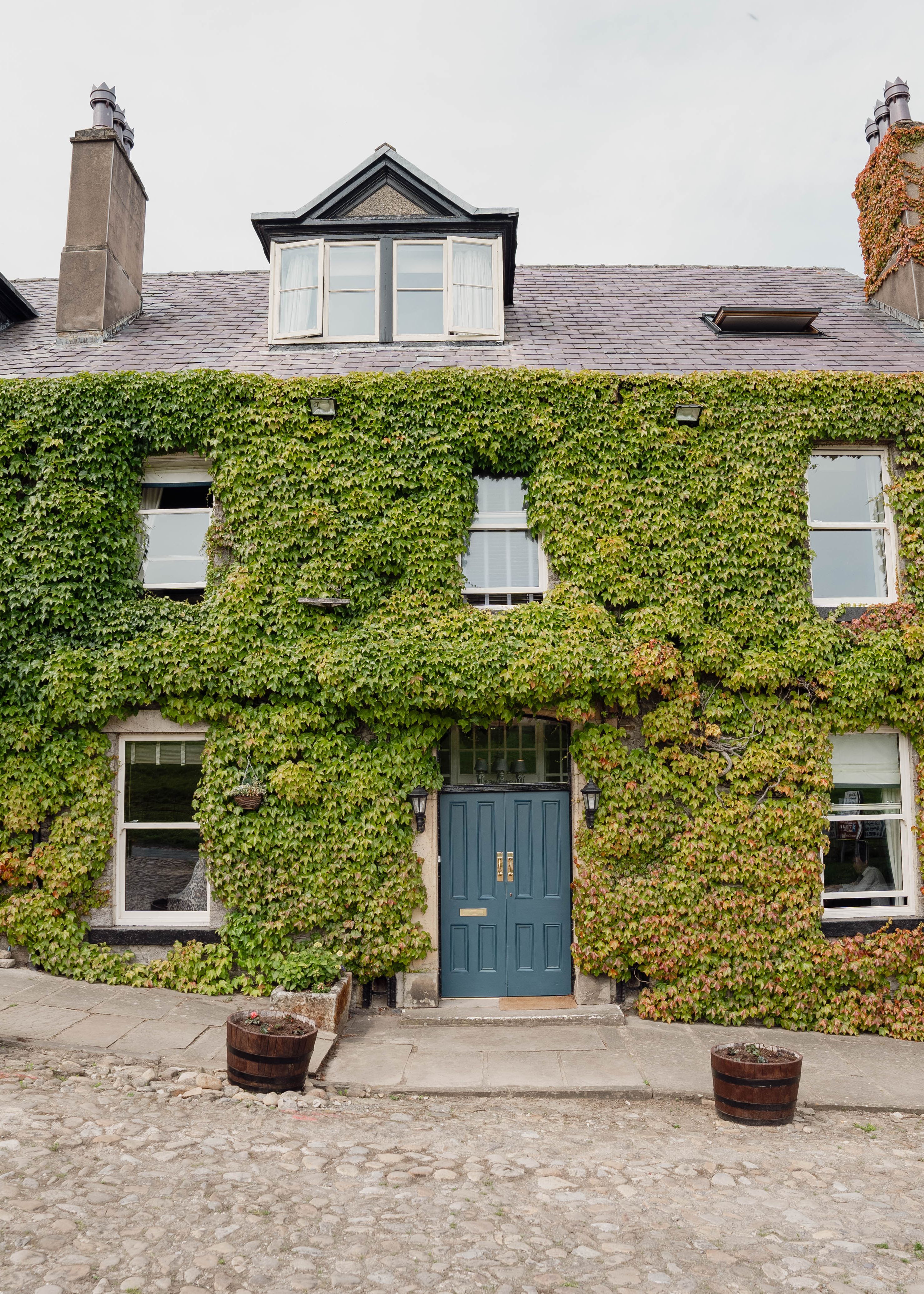 Old stone building covered in green ivy with blue doors and several windows, located on a cobblestone street