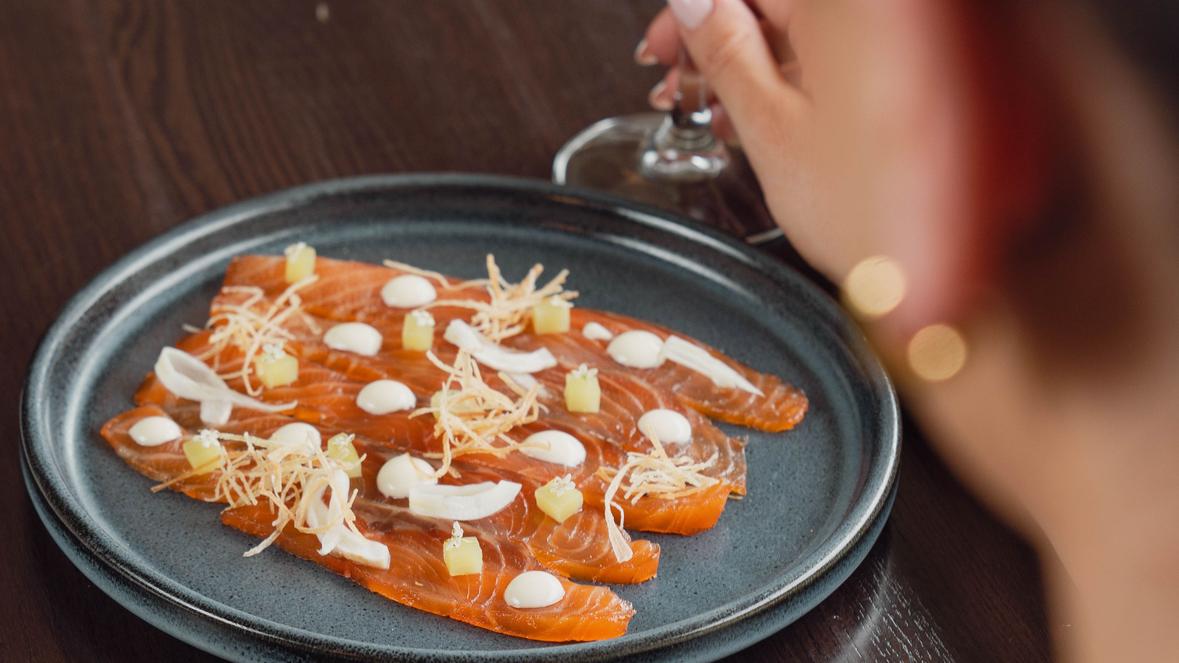 A plate of gourmet salmon sashimi with garnishes, next to a person holding a glass of white wine