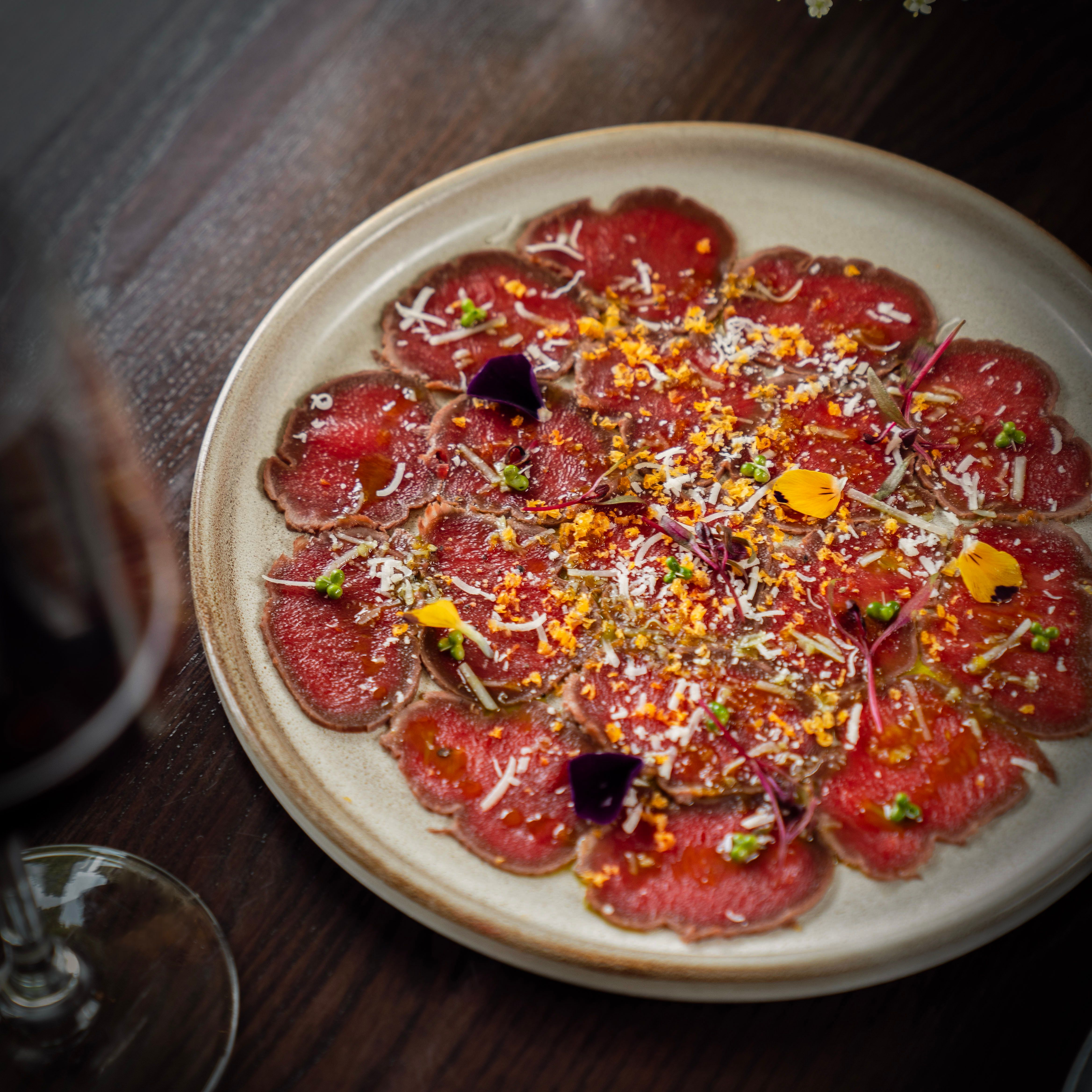 A plate of beef carpaccio garnished with edible flowers, herbs, shredded cheese, and seasoning.