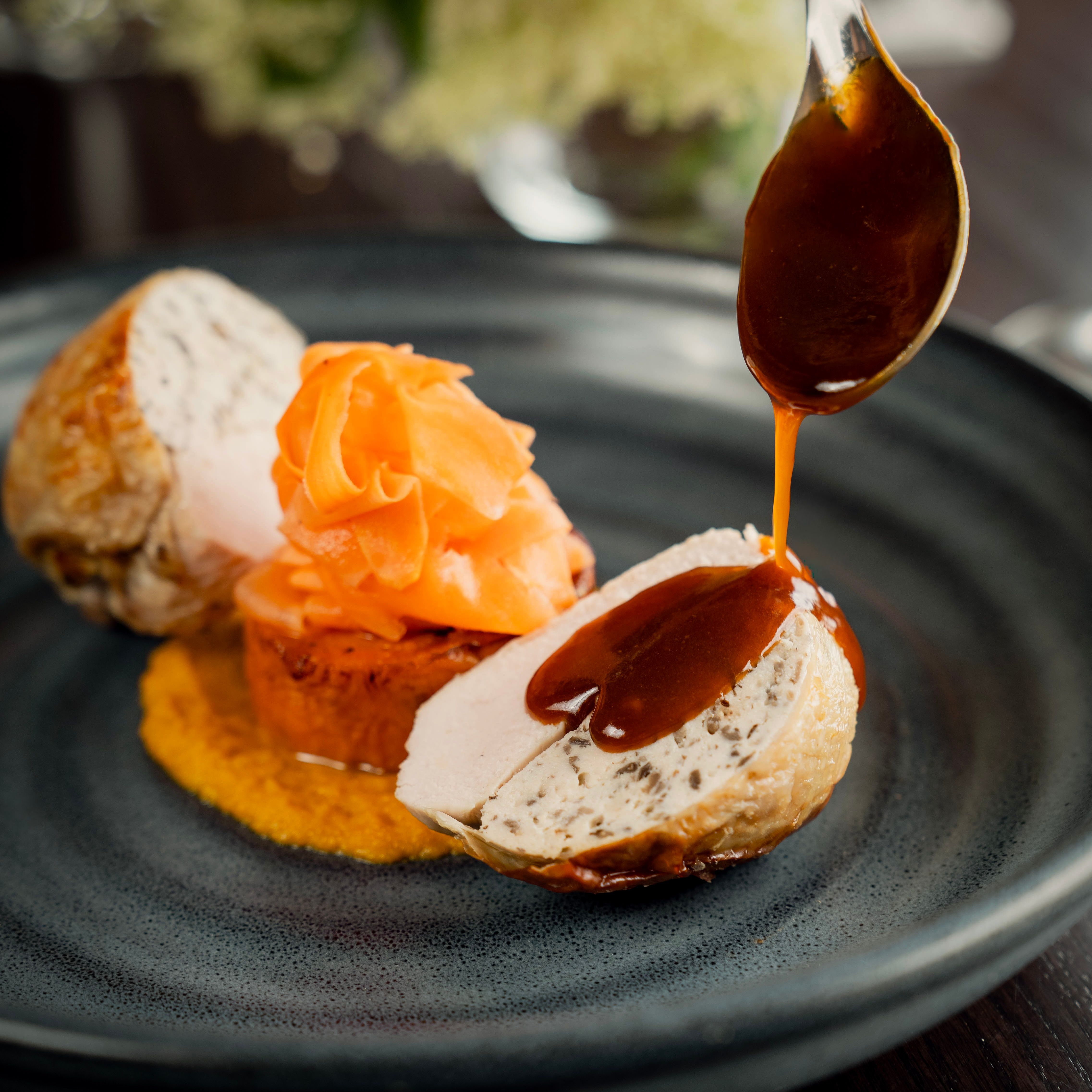 Plated gourmet chicken dish with sauce being poured over chicken breast and vegetables.