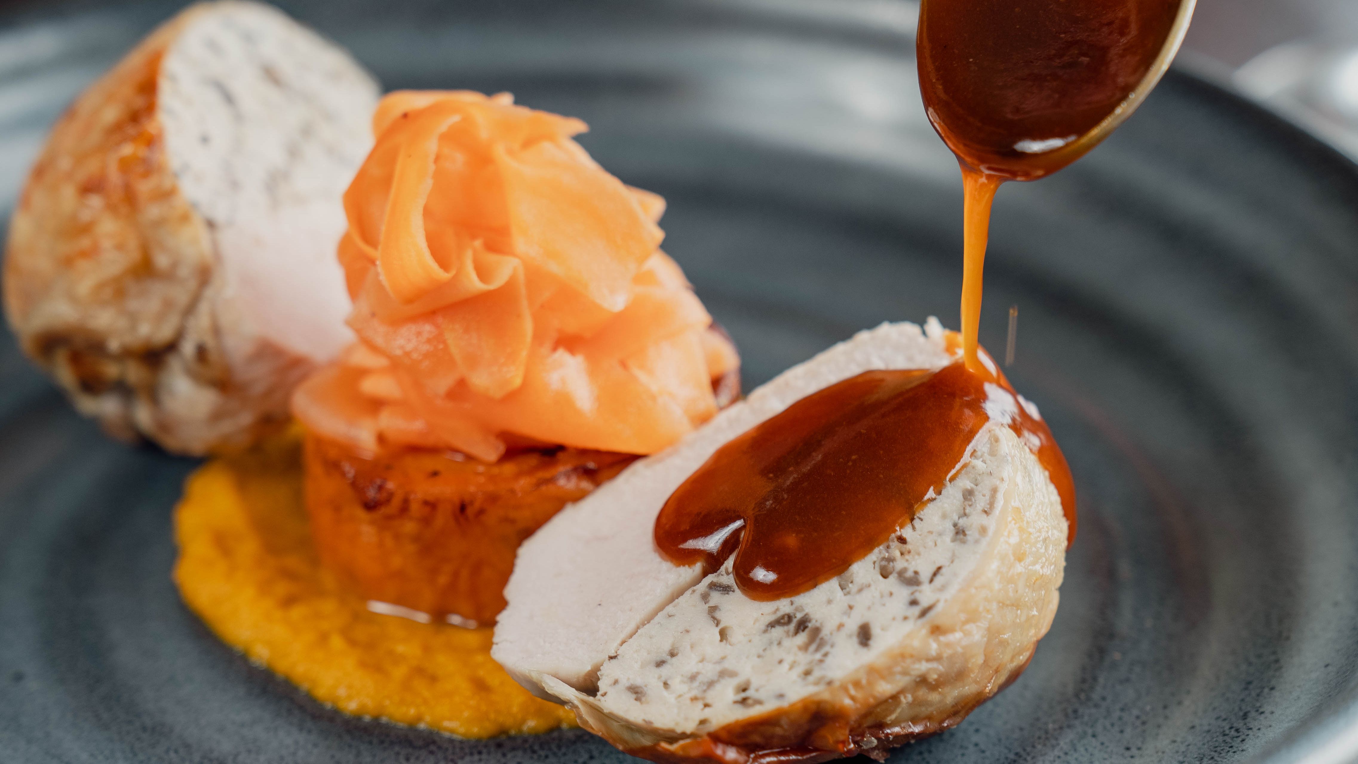 A gourmet dish with sliced chicken roulade, garnished carrots, and sauce being poured on top, served on a dark plate.