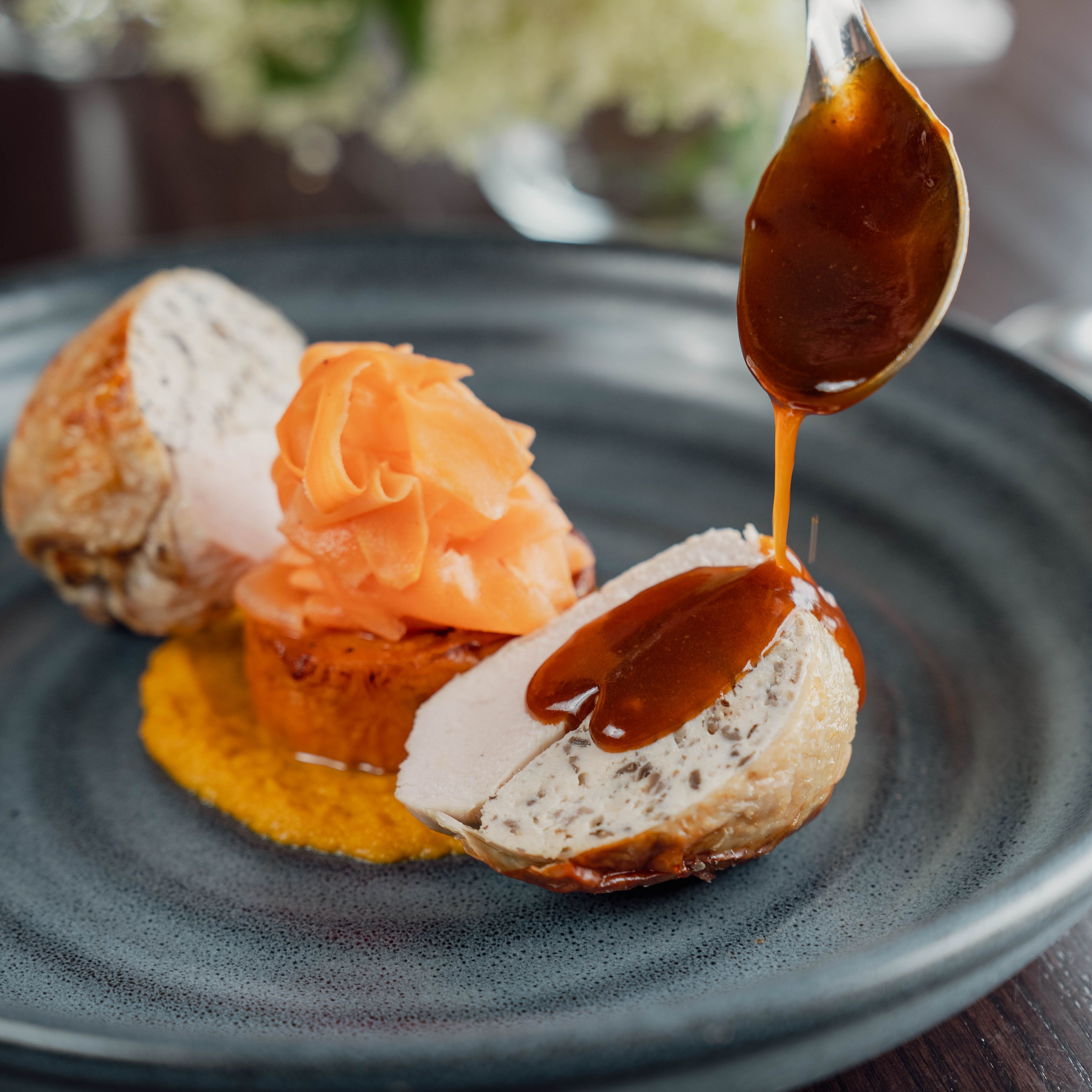 A gourmet dish with sliced chicken roulade, garnished carrots, and sauce being poured on top, served on a dark plate.