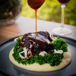 Braised beef in rich sauce on creamy mashed potatoes with greens, garnished with onions and a glass of red wine in the background.