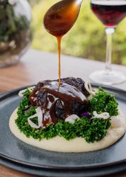 Braised beef with gravy on mashed potatoes, surrounded by greens, plated on a black dish, with a spoon pouring sauce and a glass of red wine in the background.