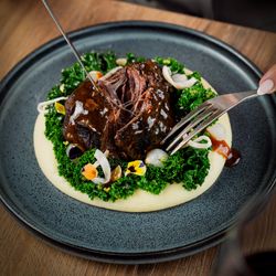 Braised beef served on mashed potatoes with kale, edible flowers, and gravy on a plate