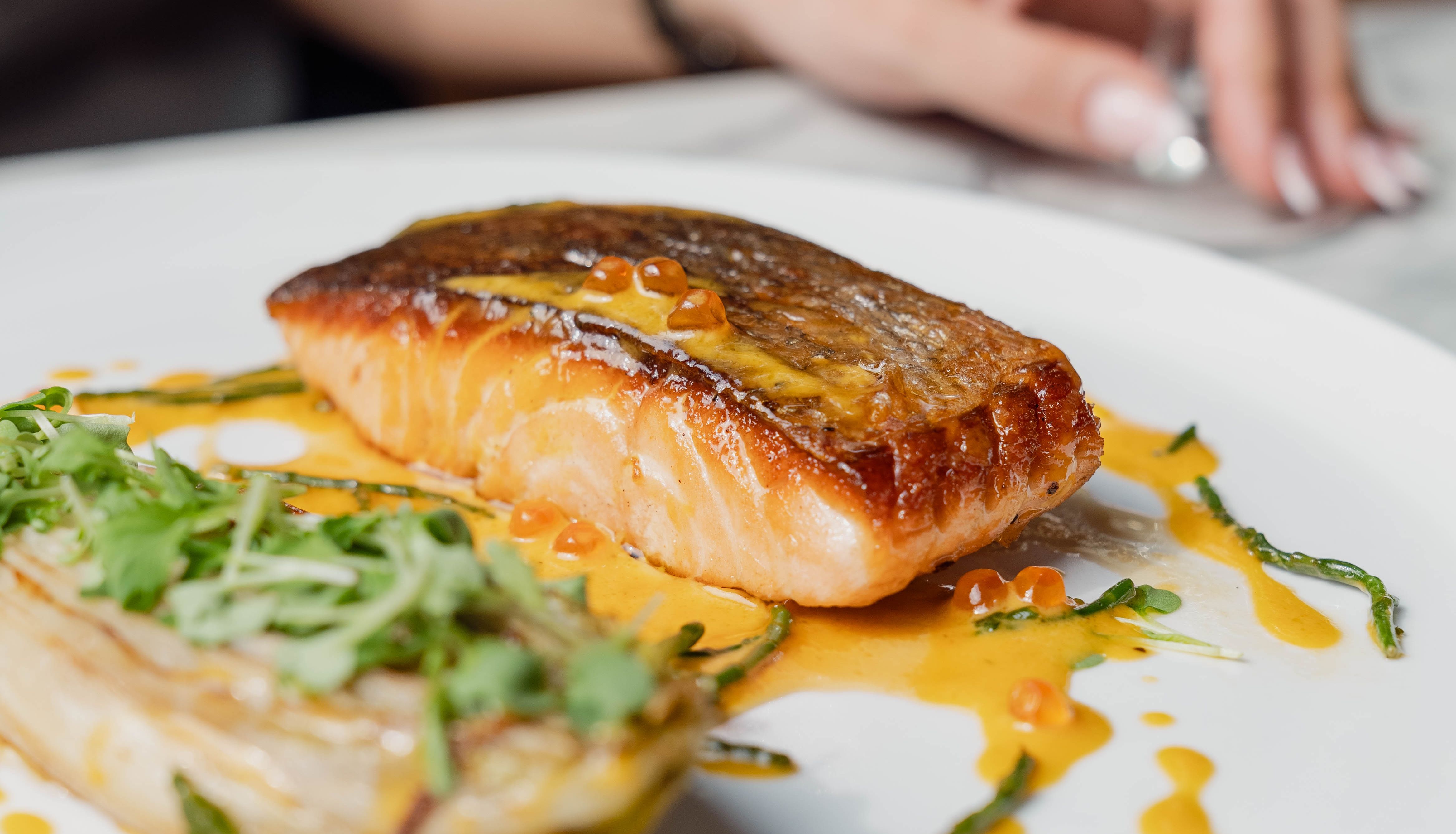 Grilled salmon fillet on a white plate with garnish and sauce, accompanied by greens. A hand and a glass of white wine appear in the background.