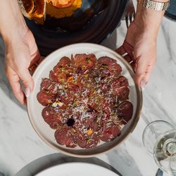 Hands holding a plate of beef carpaccio garnished with herbs and cheese, with other gourmet dishes on a marble table.