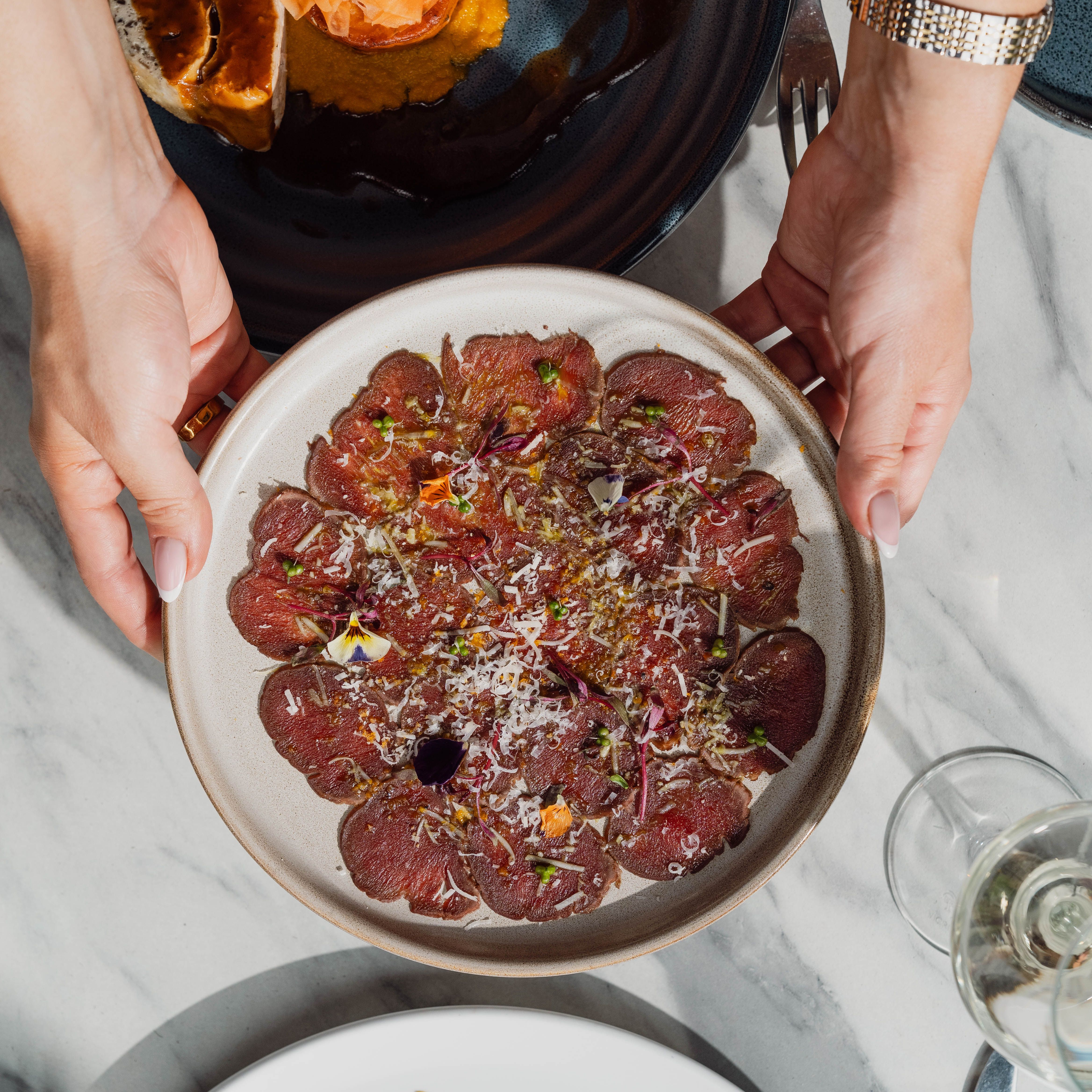 Hands holding a plate of beef carpaccio garnished with herbs and cheese, with other gourmet dishes on a marble table.