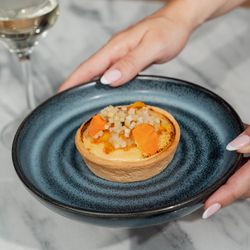 A tart dessert on a blue plate being held by a person with manicured nails, with a glass of white wine in the background.