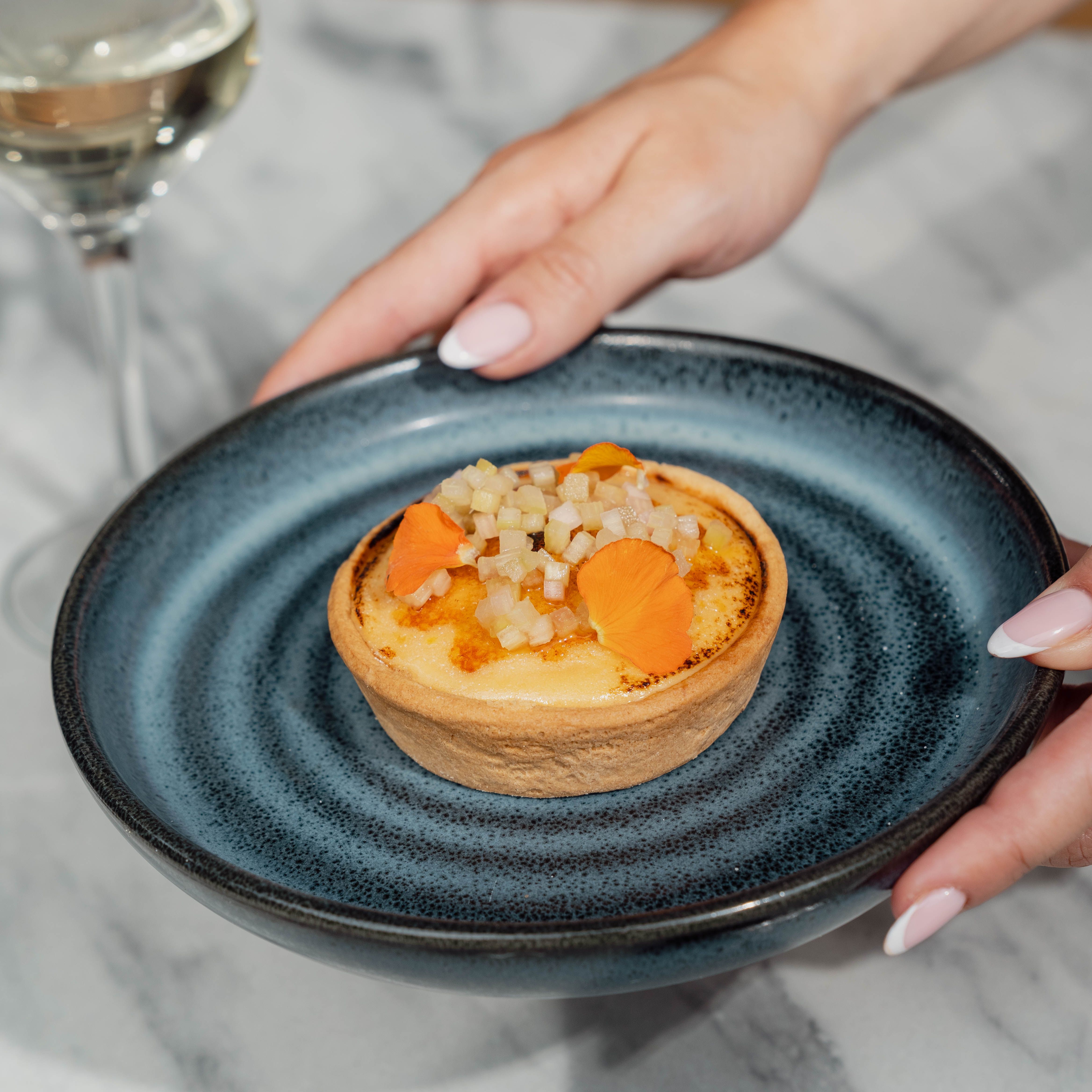 A tart dessert on a blue plate being held by a person with manicured nails, with a glass of white wine in the background.