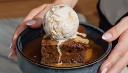A person holding a bowl with a dessert consisting of a square piece of cake topped with a scoop of ice cream and covered in caramel sauce.