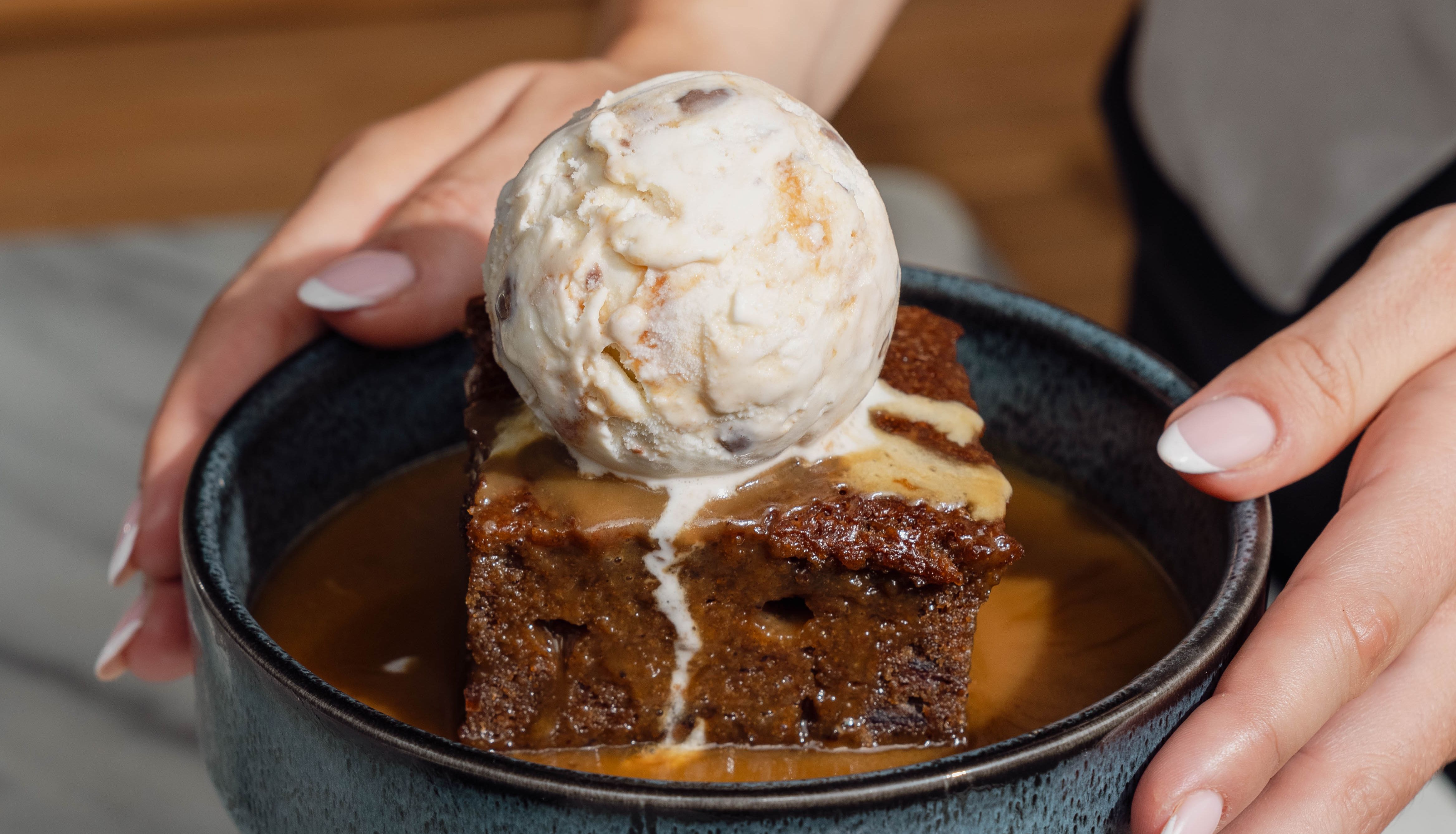 A person holding a bowl with a dessert consisting of a square piece of cake topped with a scoop of ice cream and covered in caramel sauce.