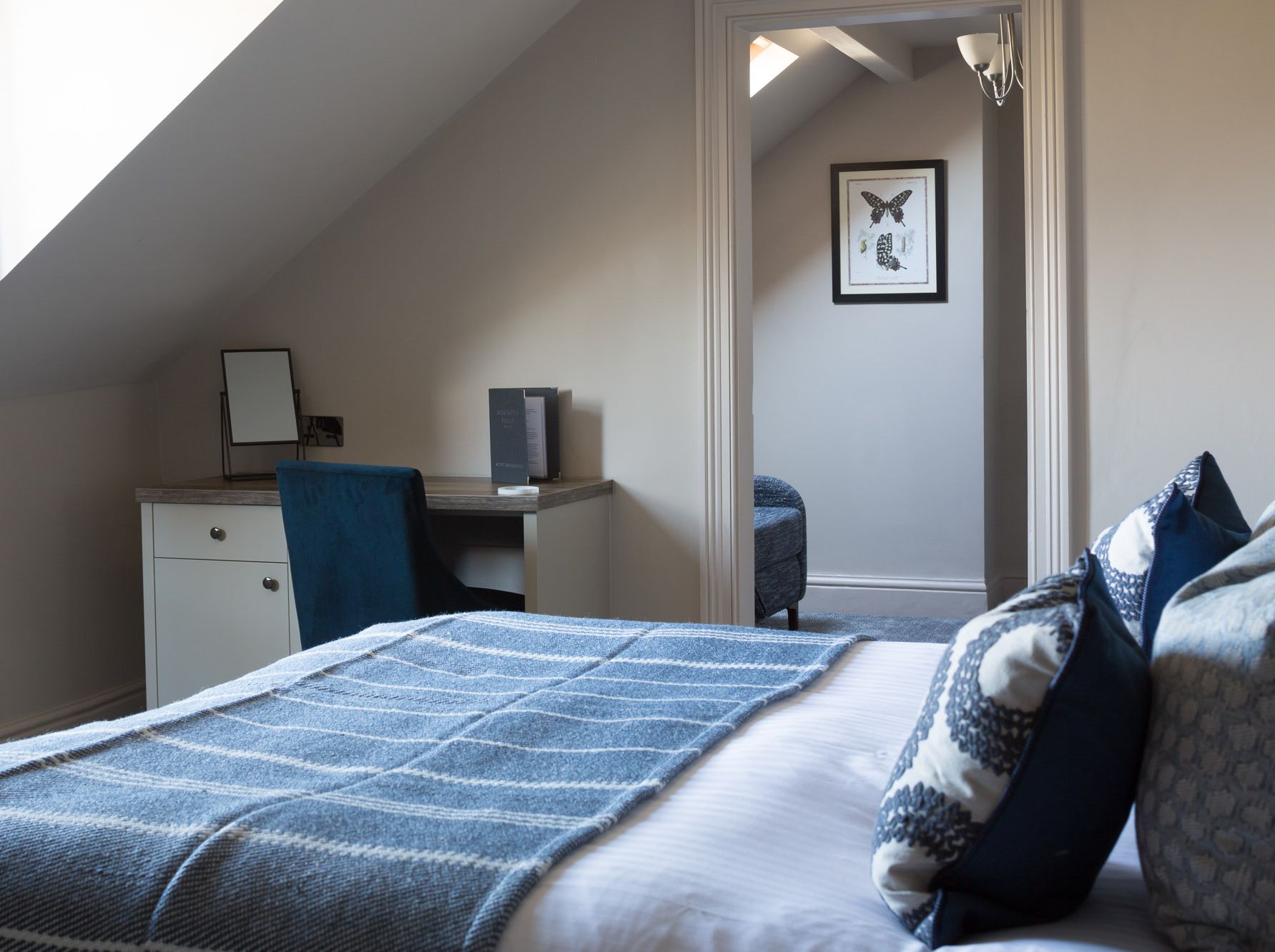 Cozy bedroom with a blue and white color scheme, a bed with patterned pillows, a desk with a blue chair, and an adjacent room with a framed butterfly print on the wall.