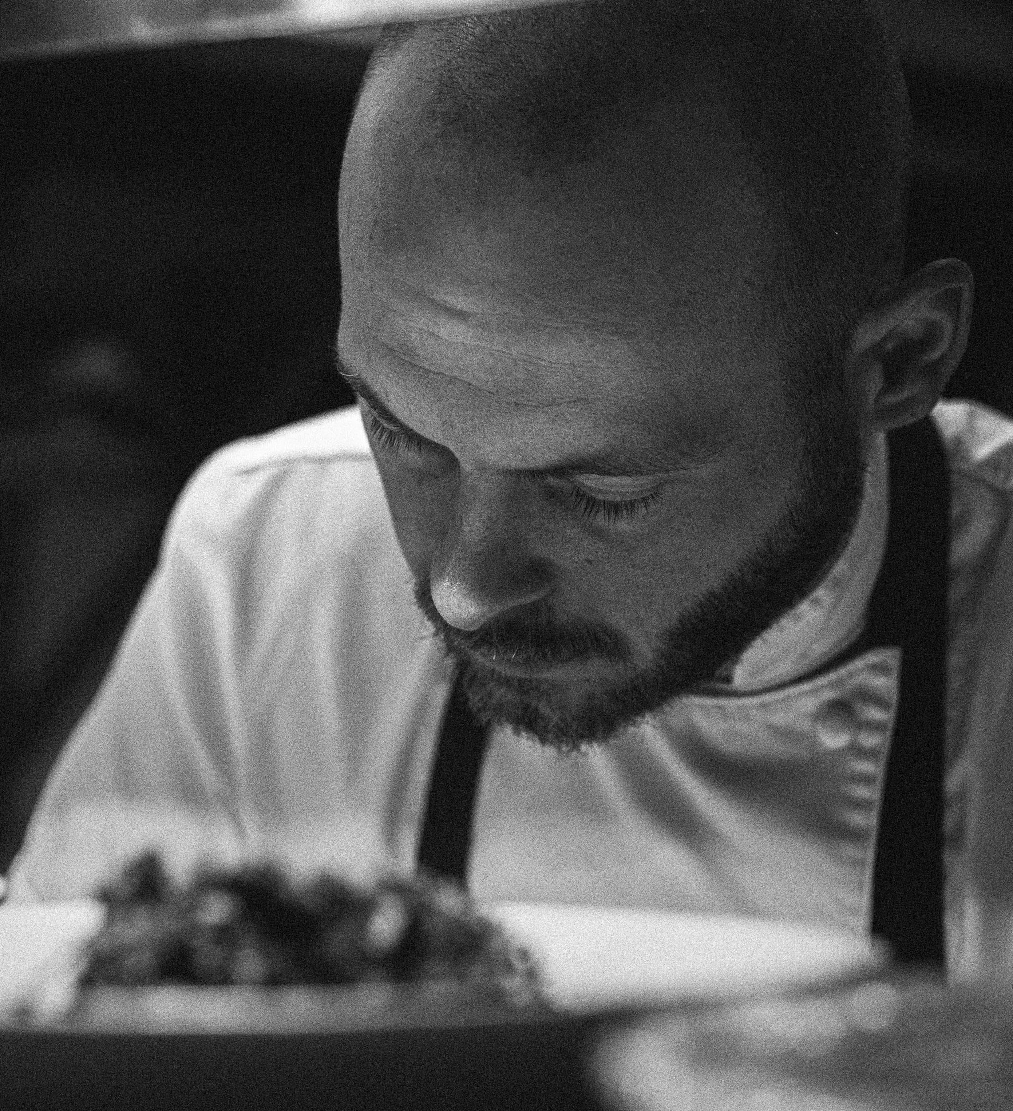Black and white photo of a young man with a beard, wearing a chef's coat and apron, looking down at a dish.