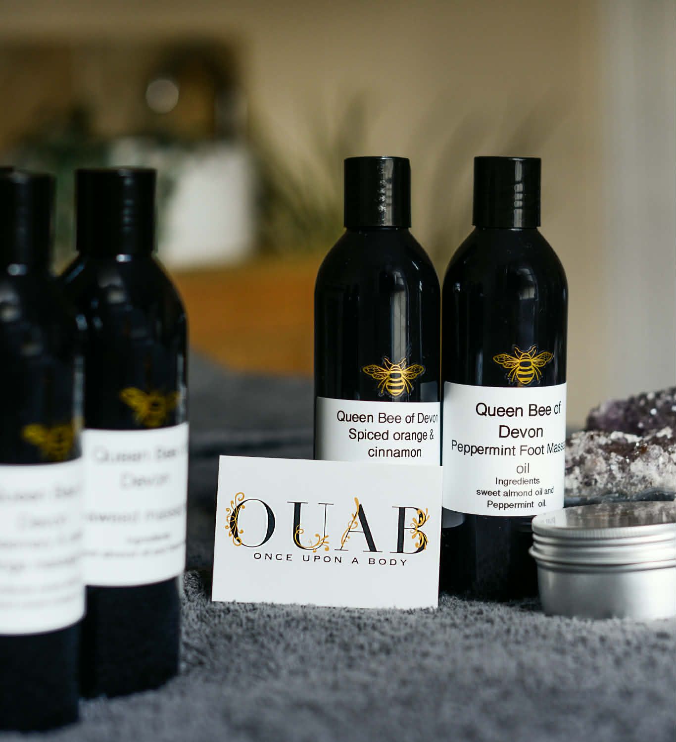 Queen Bee of Devon skincare products including Spiced orange & cinnamon and Peppermint Foot Massage oil bottles, along with a Once Upon A Body (OUAB) card on a gray towel.