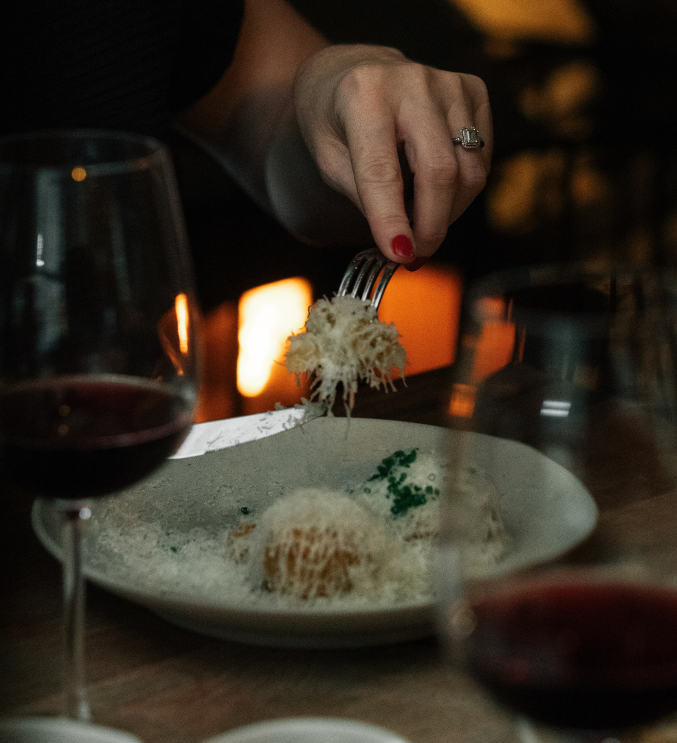 A close-up of a person holding a fork and eating a cheesy dish at a restaurant table, with two glasses of red wine in the foreground.