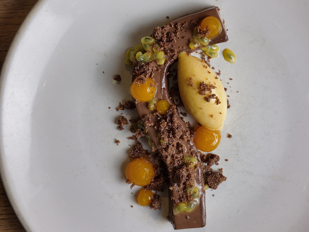Gourmet dessert with chocolate ganache, yellow fruit spheres, crumble, and sorbet on a white plate