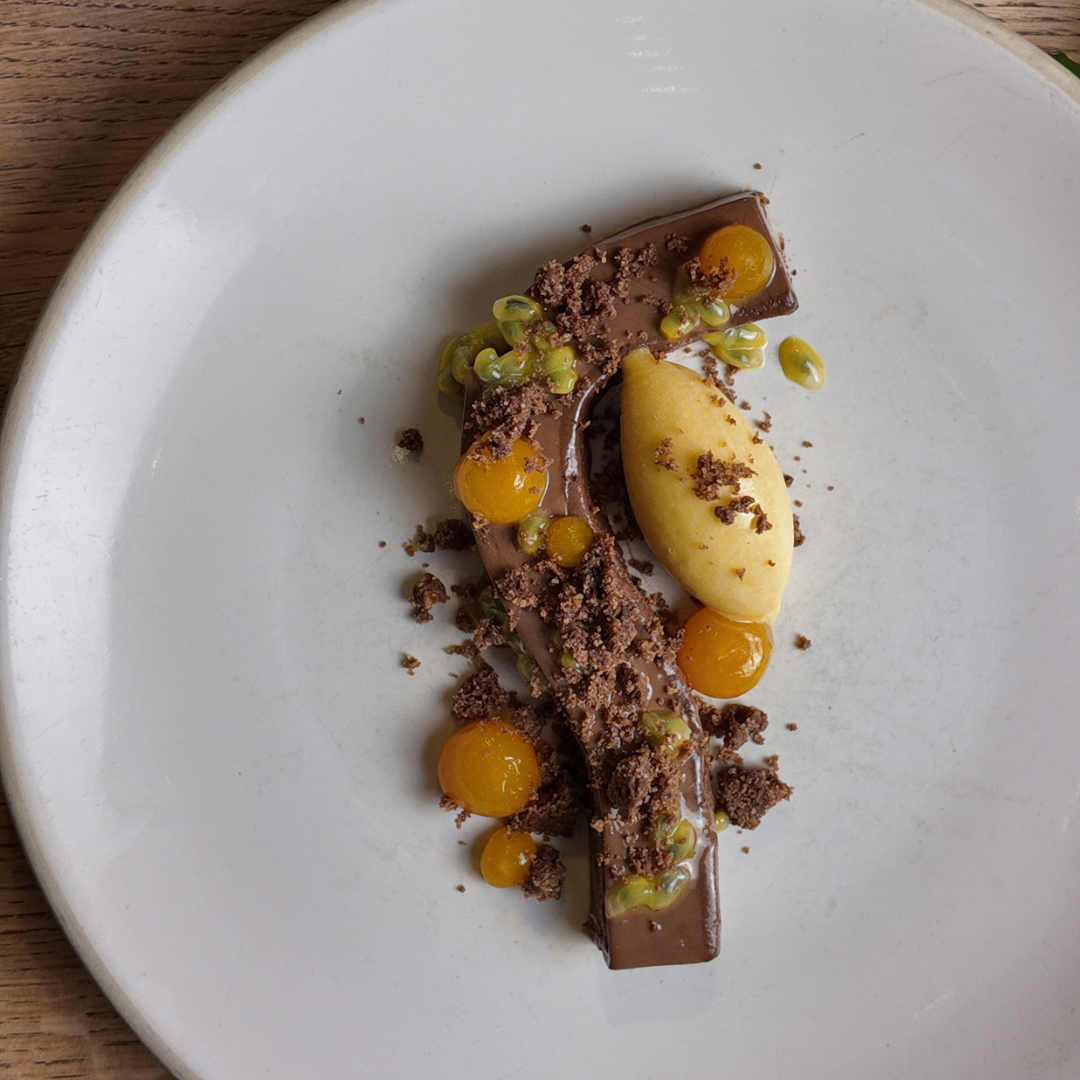 Gourmet dessert with chocolate ganache, yellow fruit spheres, crumble, and sorbet on a white plate