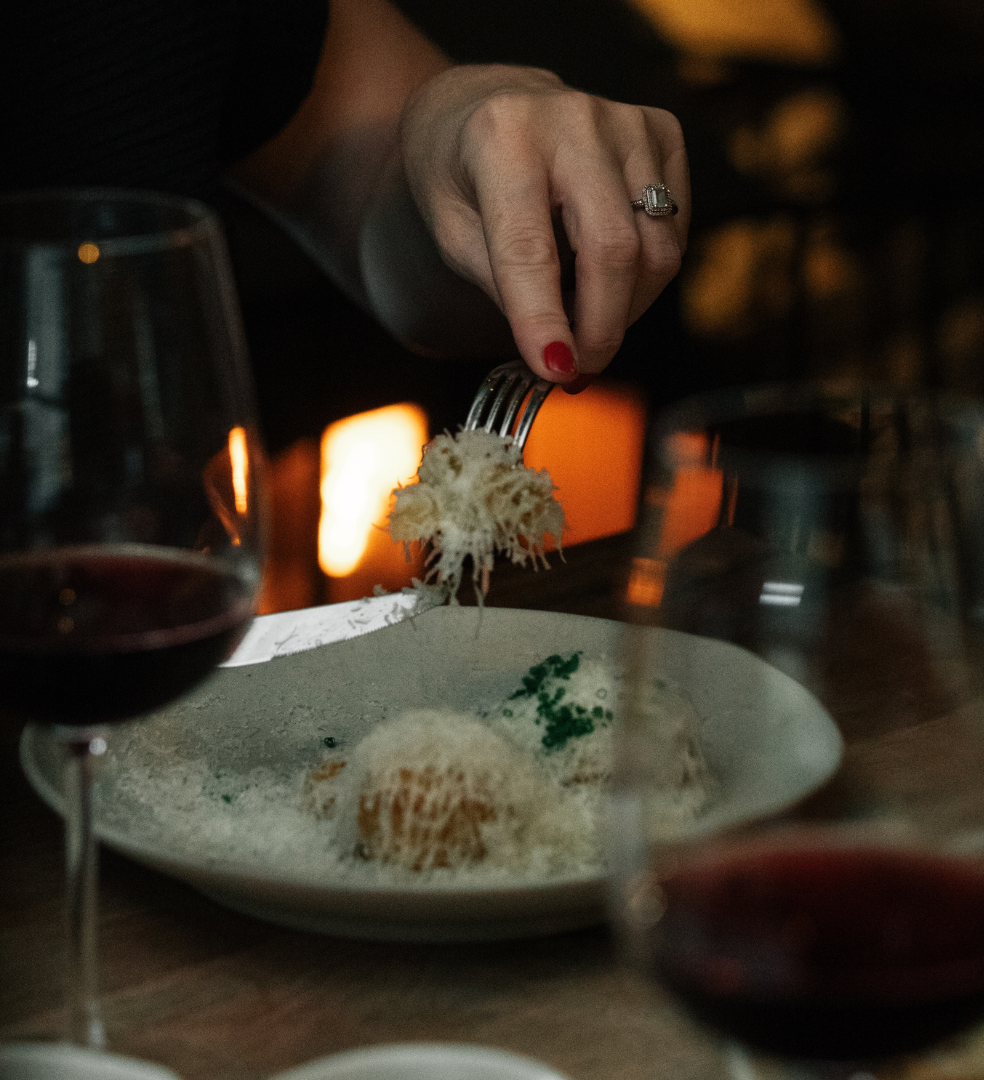 A person holding a fork with pasta topped with grated cheese, surrounded by glasses of red wine.