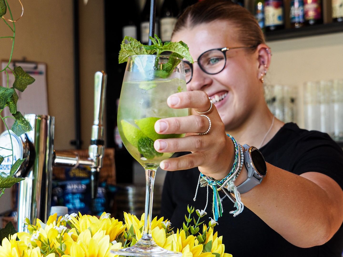 Smiling bartender holding up a cocktail garnished with mint, surrounded by yellow sunflowers.
