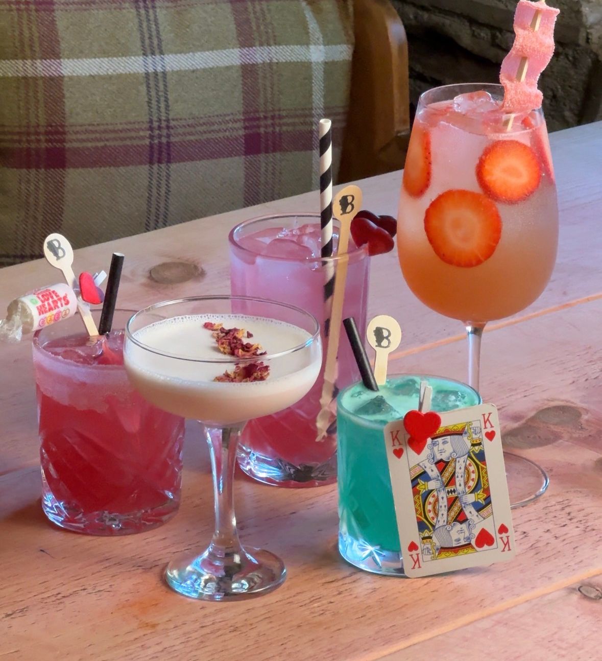 A selection of colorful cocktails garnished with fruit, candy, and a playing card, displayed on a rustic wooden table.