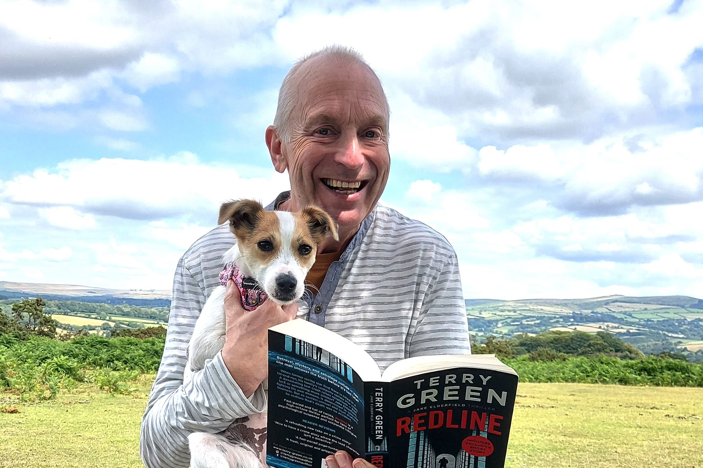 Man sitting on a bench in a scenic outdoor setting, smiling while holding a small dog and reading a book titled 'Redline' by Terry Green.
