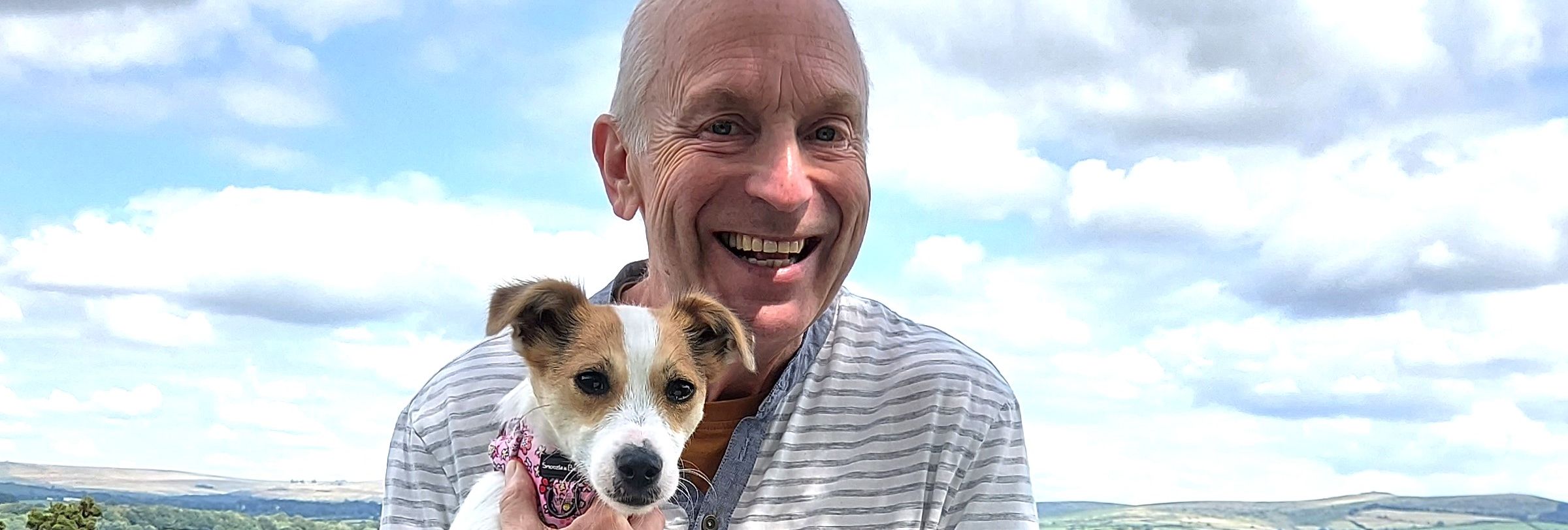 Man sitting on a bench in a scenic outdoor setting, smiling while holding a small dog and reading a book titled 'Redline' by Terry Green.