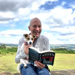 Man sitting on a bench in a scenic outdoor setting, smiling while holding a small dog and reading a book titled 'Redline' by Terry Green.
