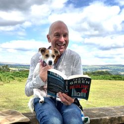 Man sitting on a bench in a scenic outdoor setting, smiling while holding a small dog and reading a book titled 'Redline' by Terry Green.