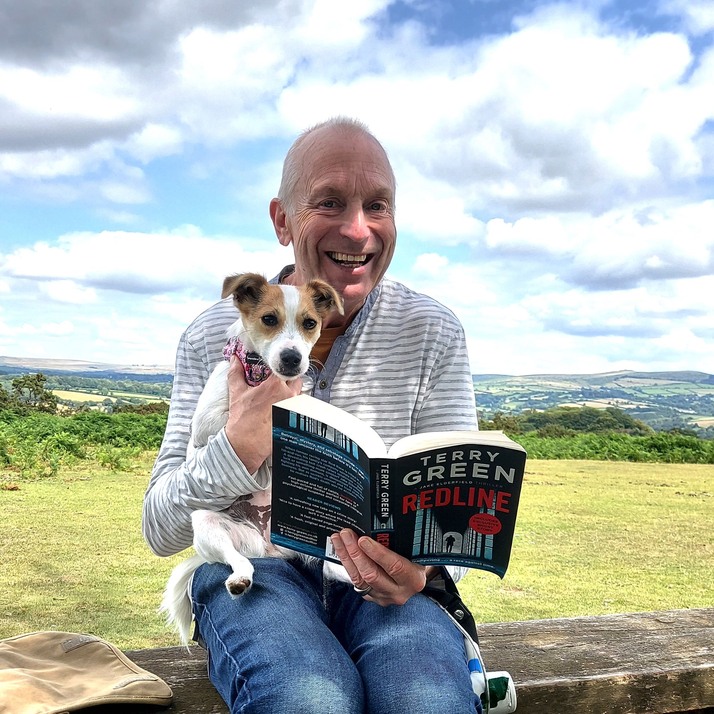 Man sitting on a bench in a scenic outdoor setting, smiling while holding a small dog and reading a book titled 'Redline' by Terry Green.